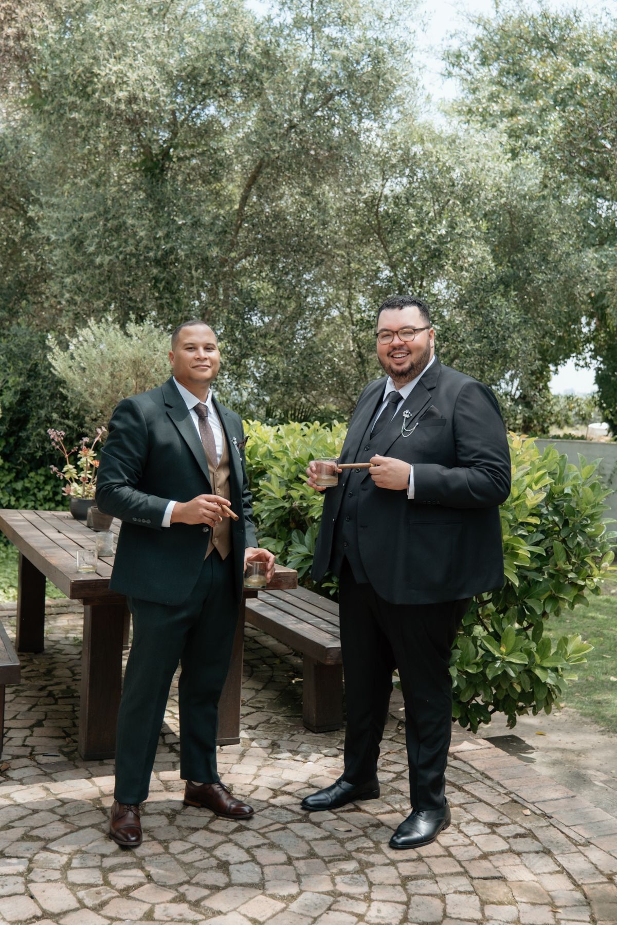 Two groomsmen in dark suits pose together on a cobblestone patio surrounded by lush greenery.