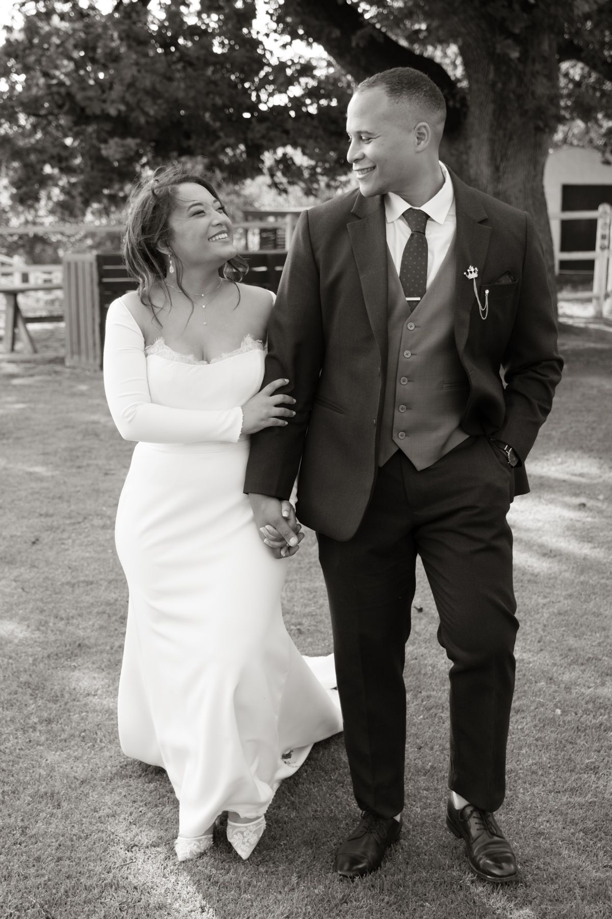 Black and white photo of smiling bride and groom walking hand in hand at outdoor wedding.