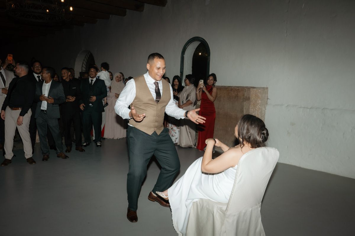 Energetic wedding reception dance floor moment with groom in vest reaching toward bride in white dress.