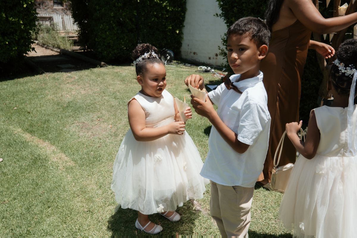 Two children in formal white attire playing with bubbles on green grass at an outdoor wedding celebration.