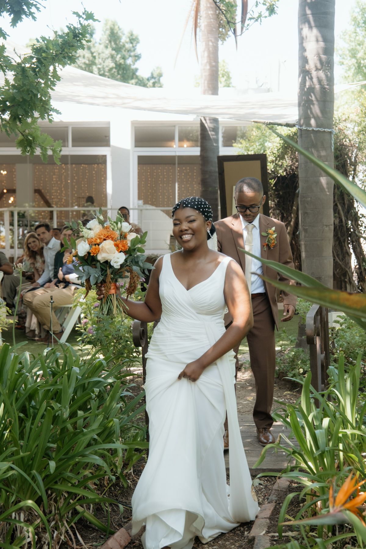 A radiant bride in a white gown holds a bouquet while walking down an outdoor garden wedding aisle.