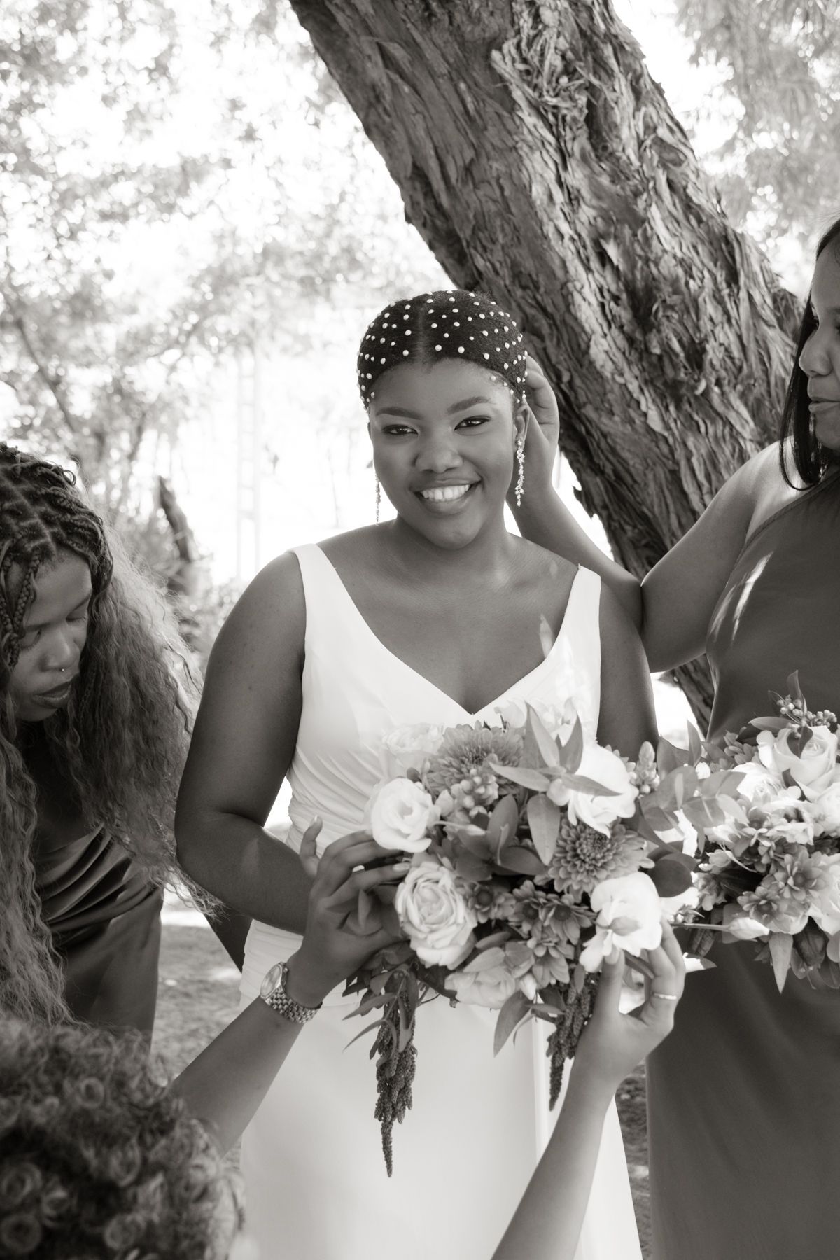 Black and white photo of smiling bride in white gown holding floral bouquet surrounded by bridesmaids under trees.