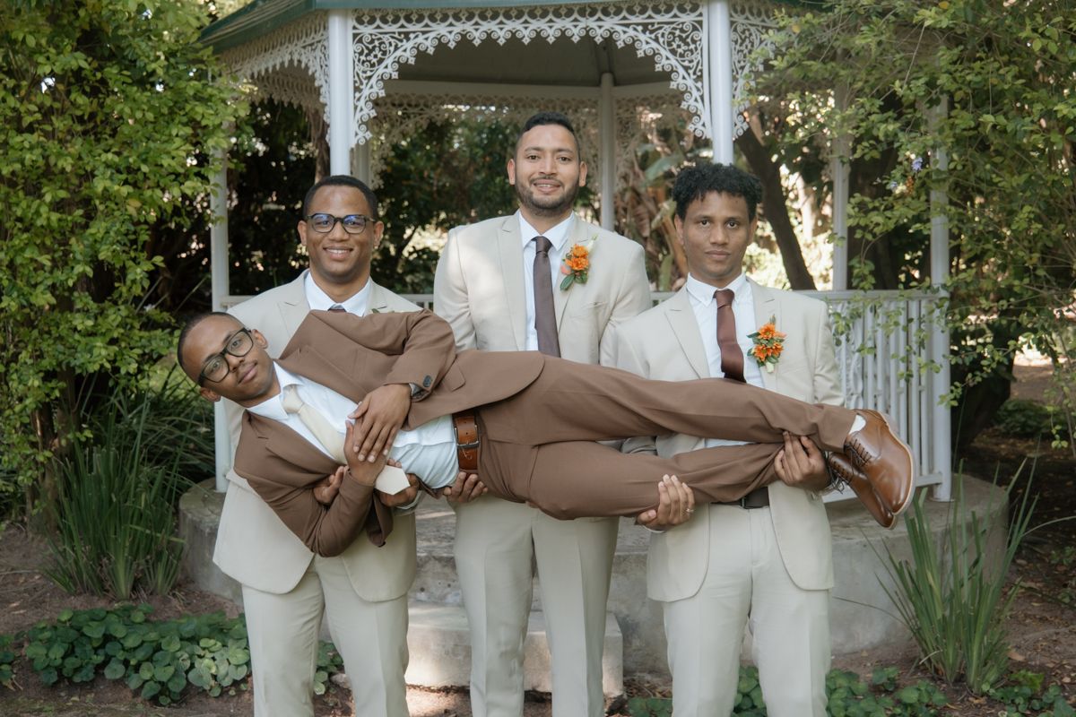 Four groomsmen pose humorously, lifting one man horizontally in front of a white gazebo.