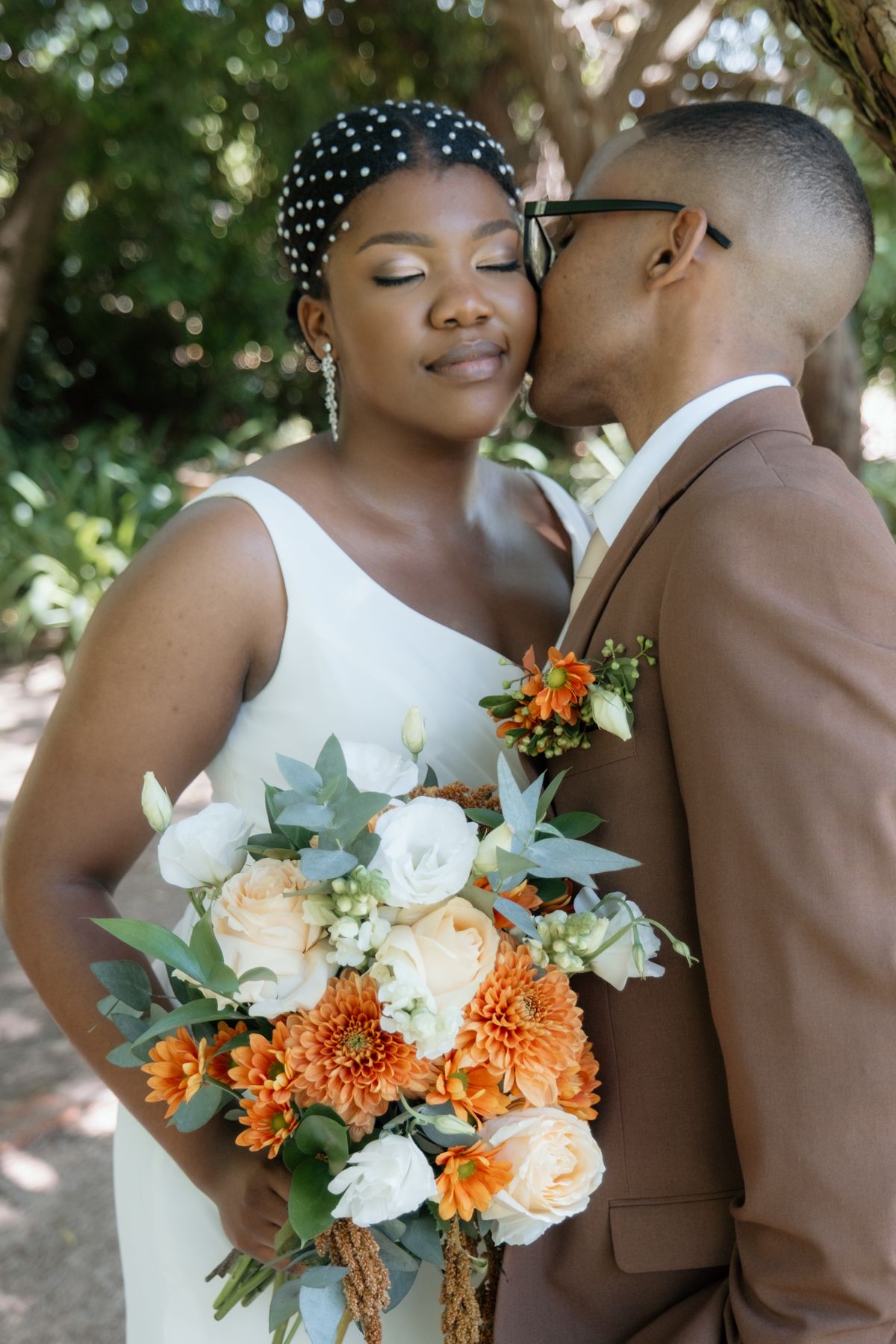 Groom in brown suit kissing bride's cheek as she holds a vibrant orange and white wedding bouquet.