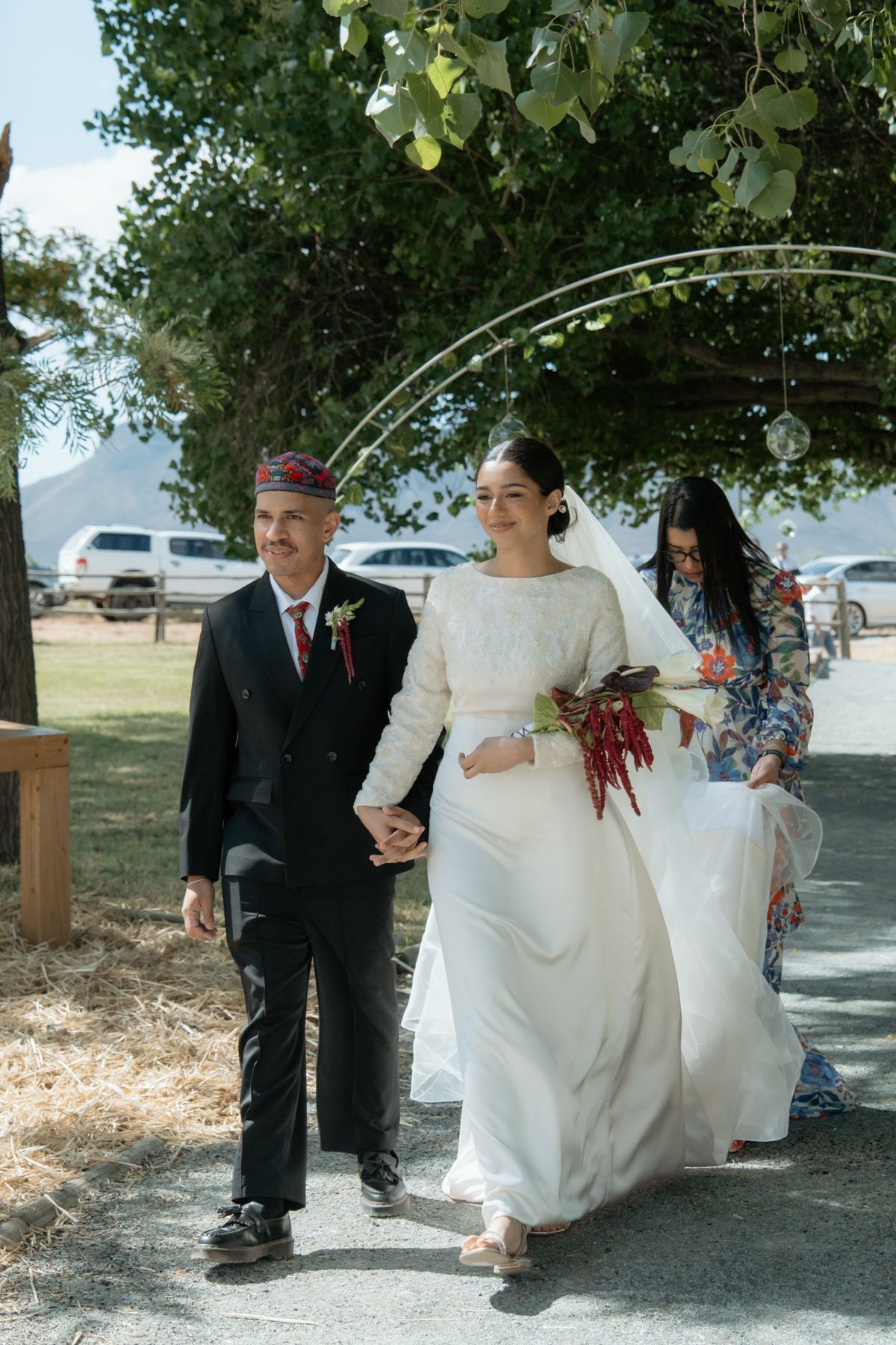 Bride in white lace gown walks with groom in black suit and red cap at outdoor wedding ceremony.