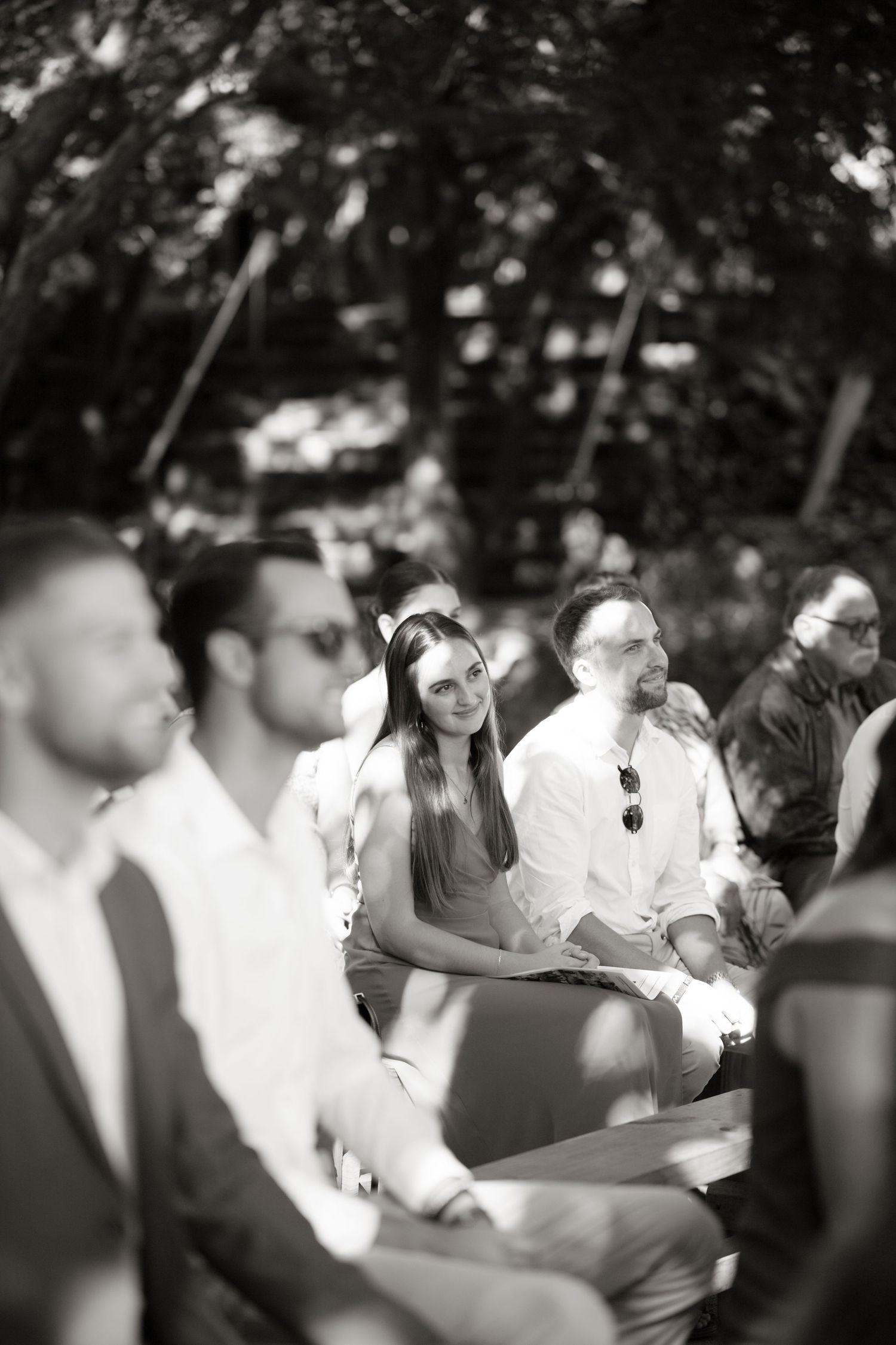 Black and white photo of smiling wedding guests seated outdoors during a garden wedding ceremony.