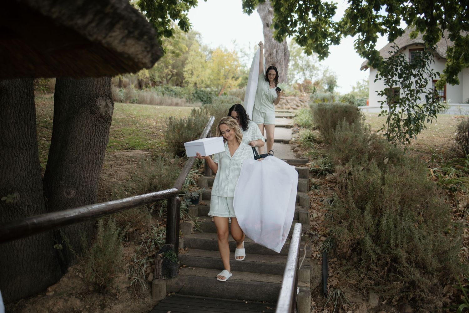 Two bridesmaids in mint dresses carry white wedding items down outdoor stairs surrounded by lush greenery.