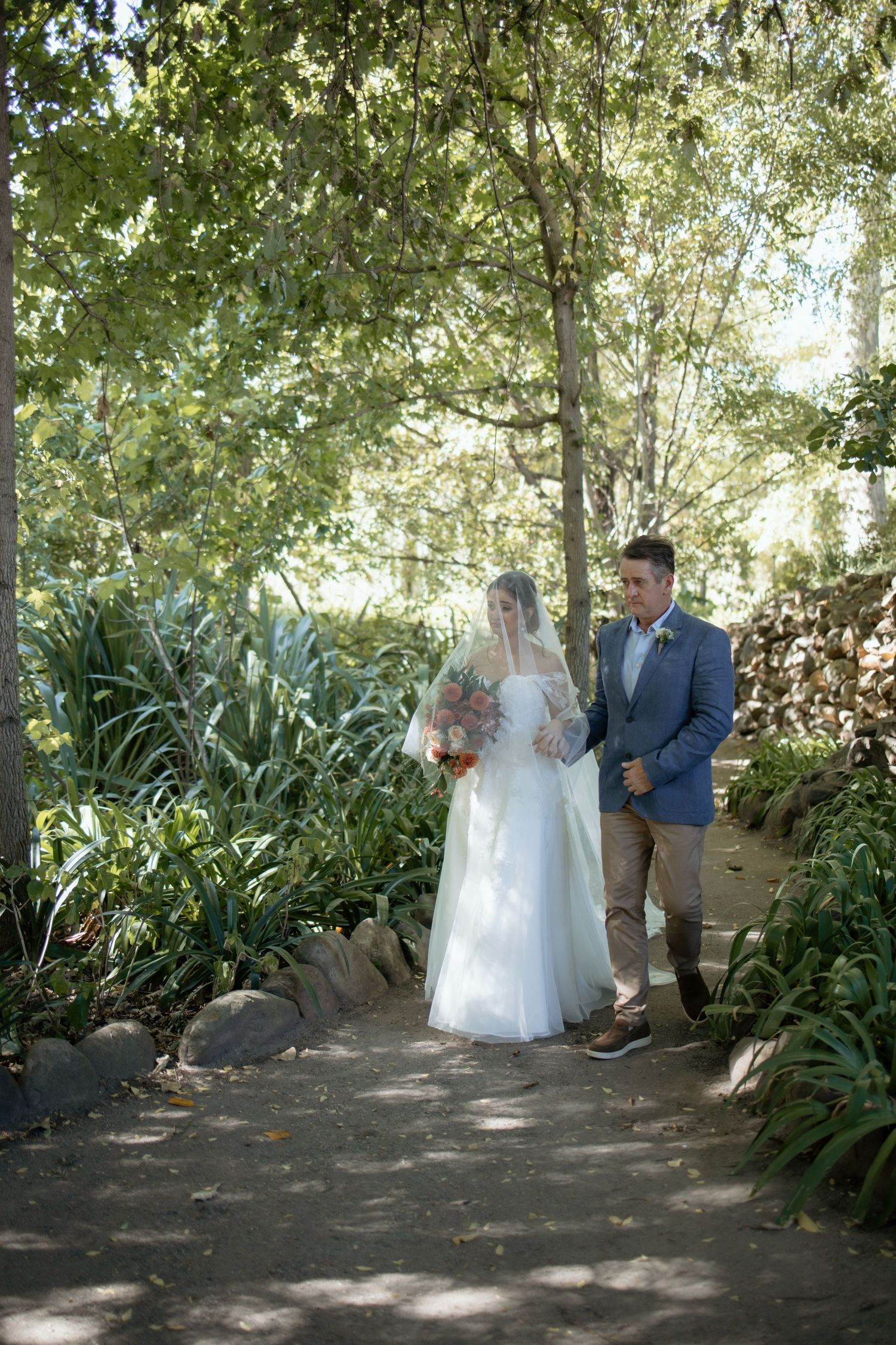 Bride in white gown with veil and groom in blue suit walk together along a lush garden pathway.