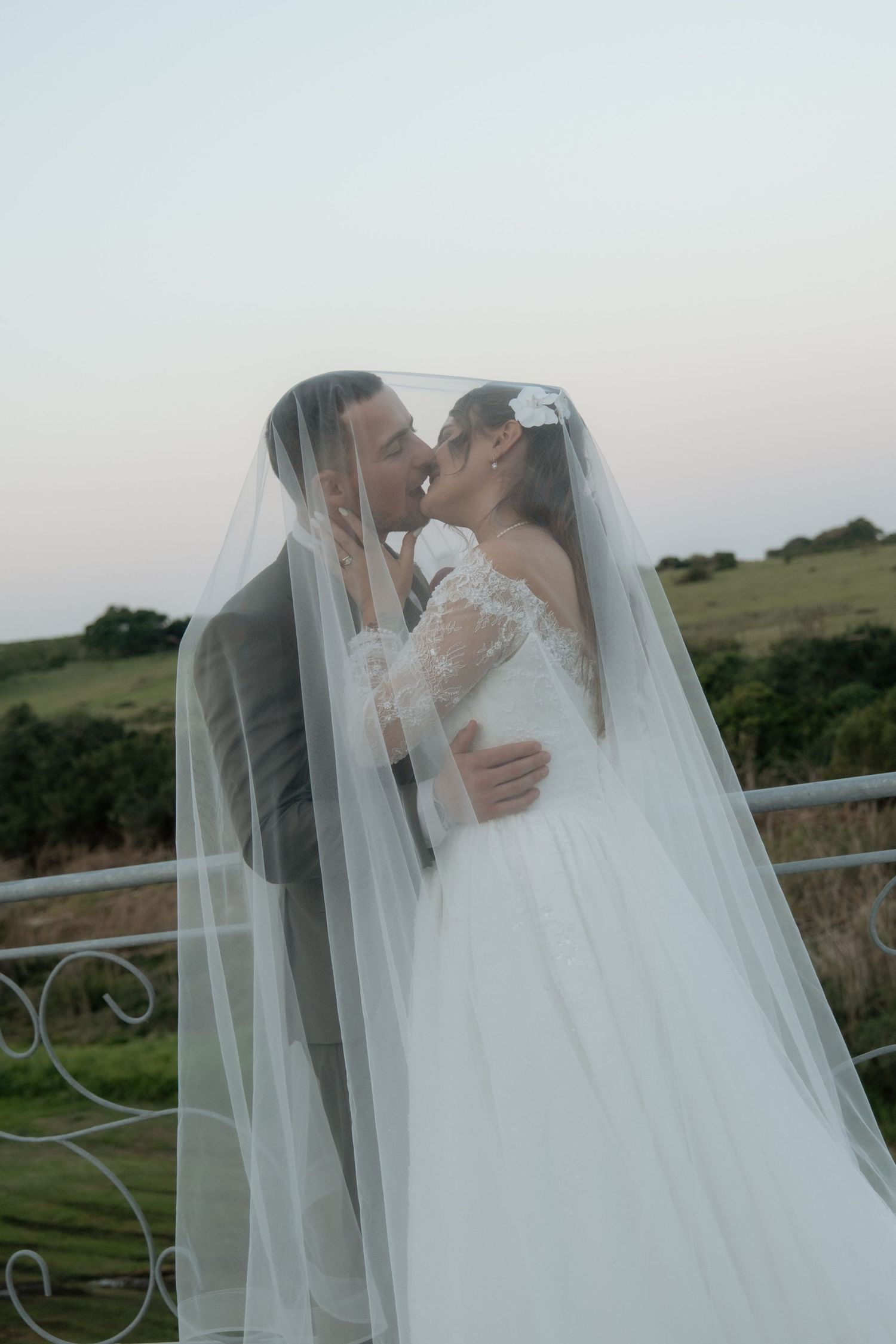 Bride and groom sharing a kiss under flowing white veil with green hills behind them.