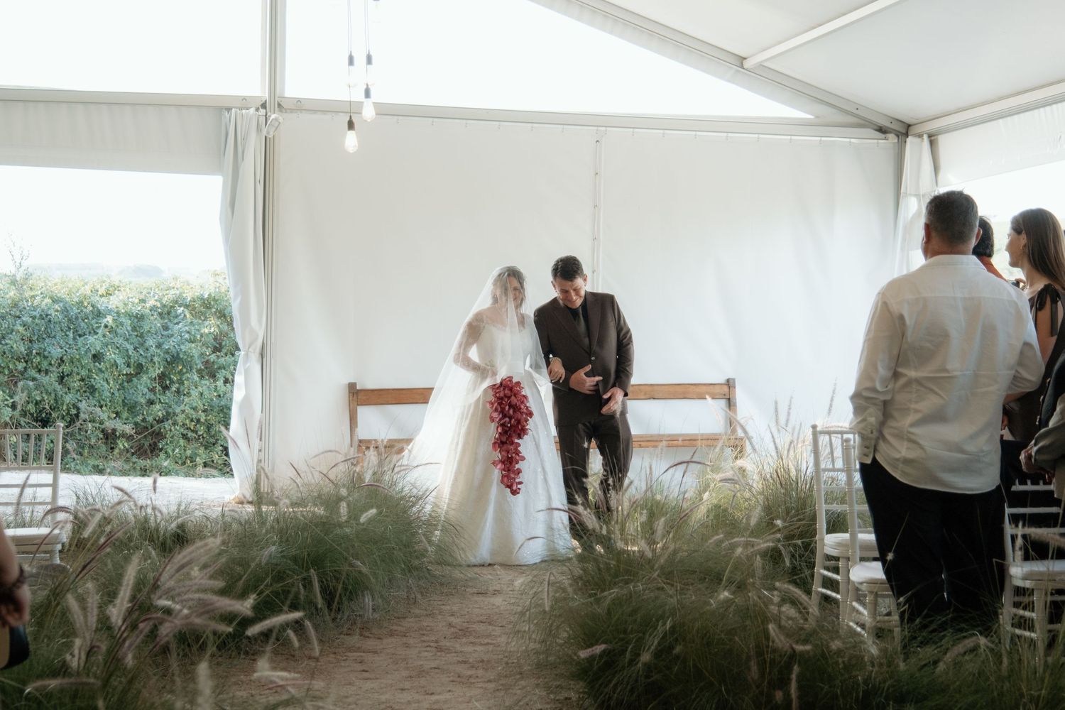 Bride and groom stand at outdoor wedding altar under white tent, surrounded by tall grass and white chairs.