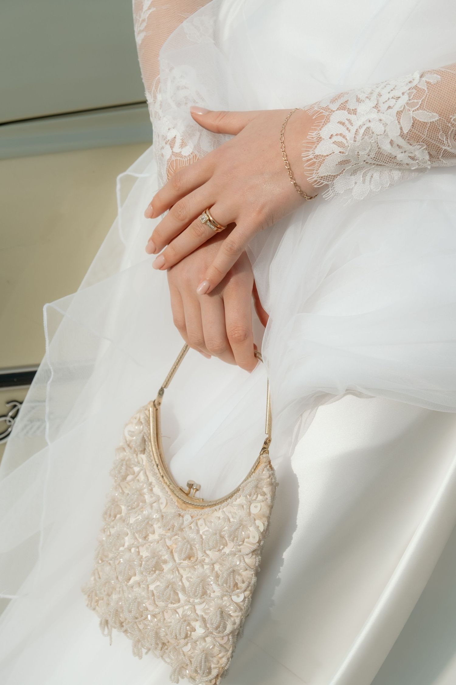 Close-up of bride's hands holding beaded ivory clutch purse, wearing gold ring and bracelet.