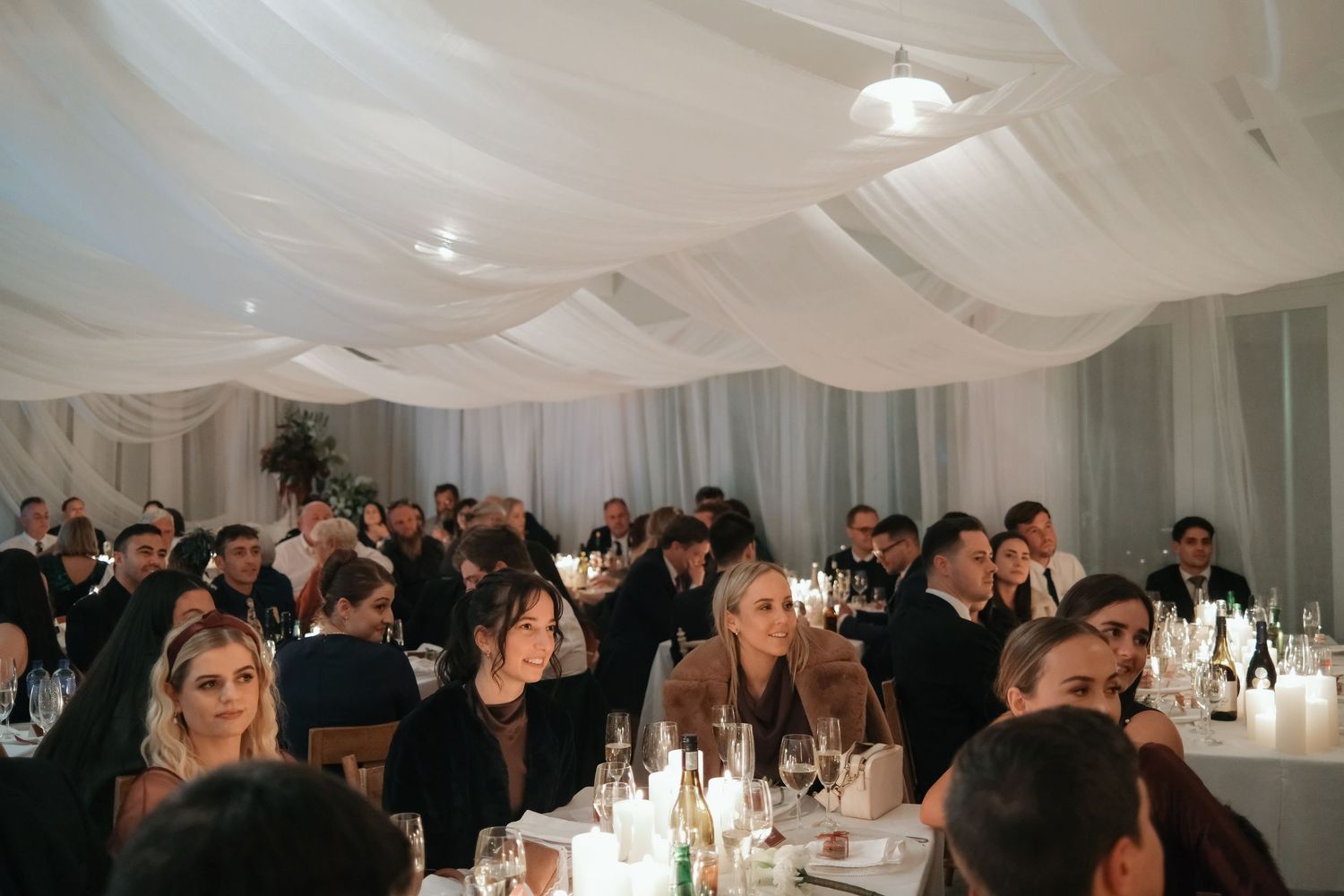Elegant wedding reception in a white draped marquee tent with guests seated at candlelit tables.