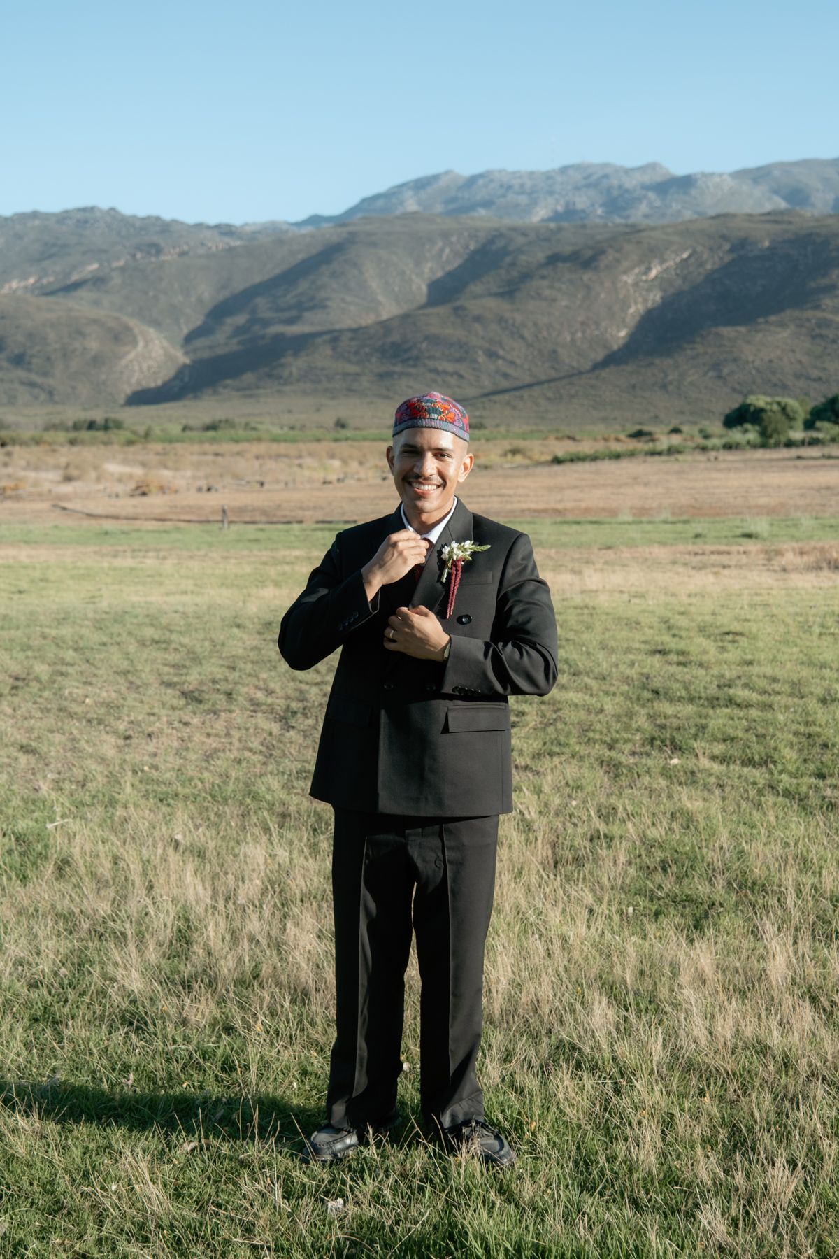 Smiling groom in black suit and traditional cap stands in open field with mountain landscape behind.