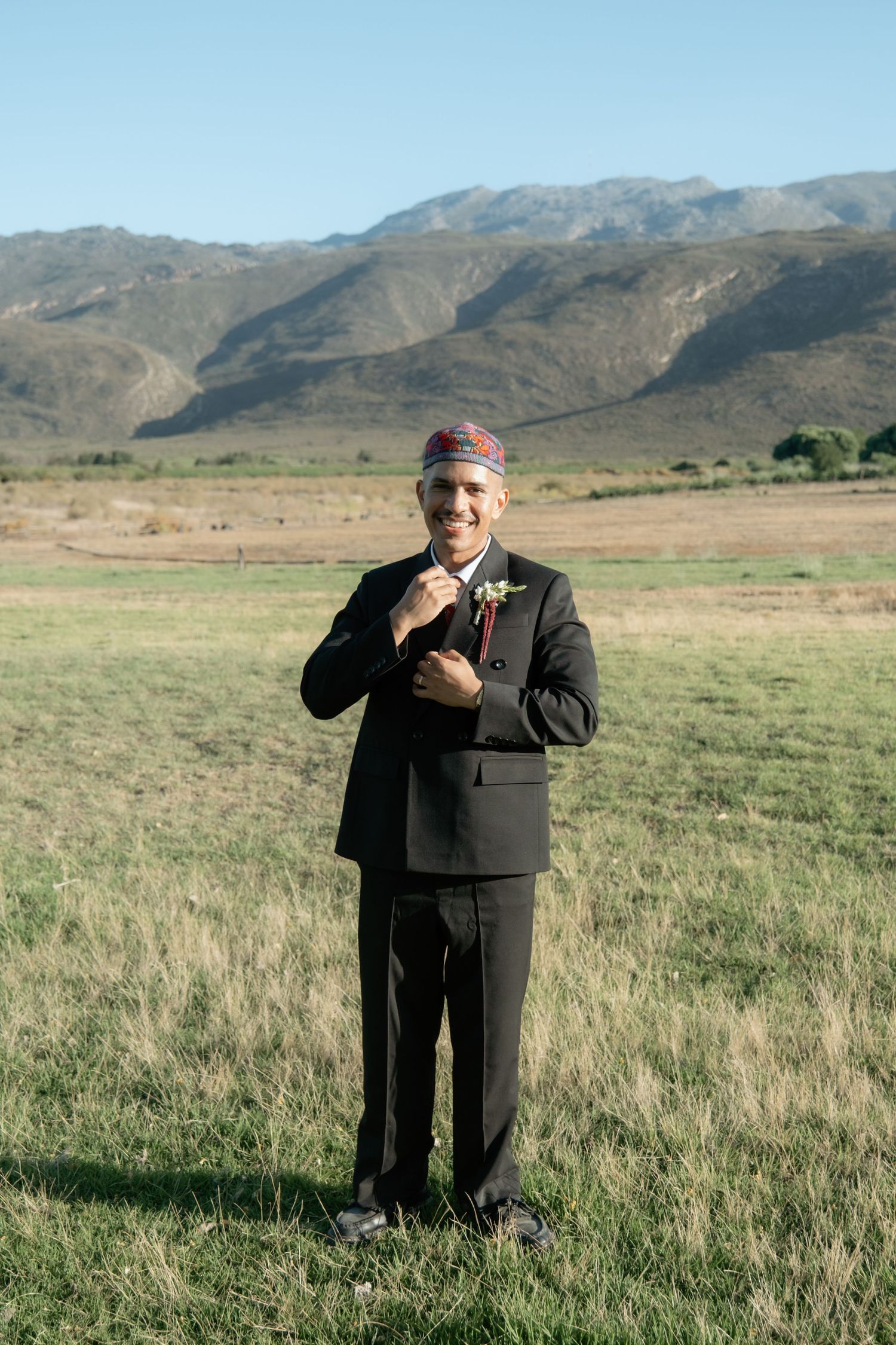 Smiling groom in black suit and traditional red kufi cap adjusting boutonniere in scenic mountain field.