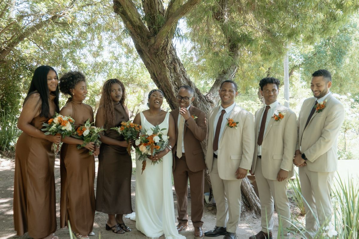 Wedding party poses under a tree, bride in white with bridesmaids in brown dresses and groomsmen in tan suits holding bouquets.