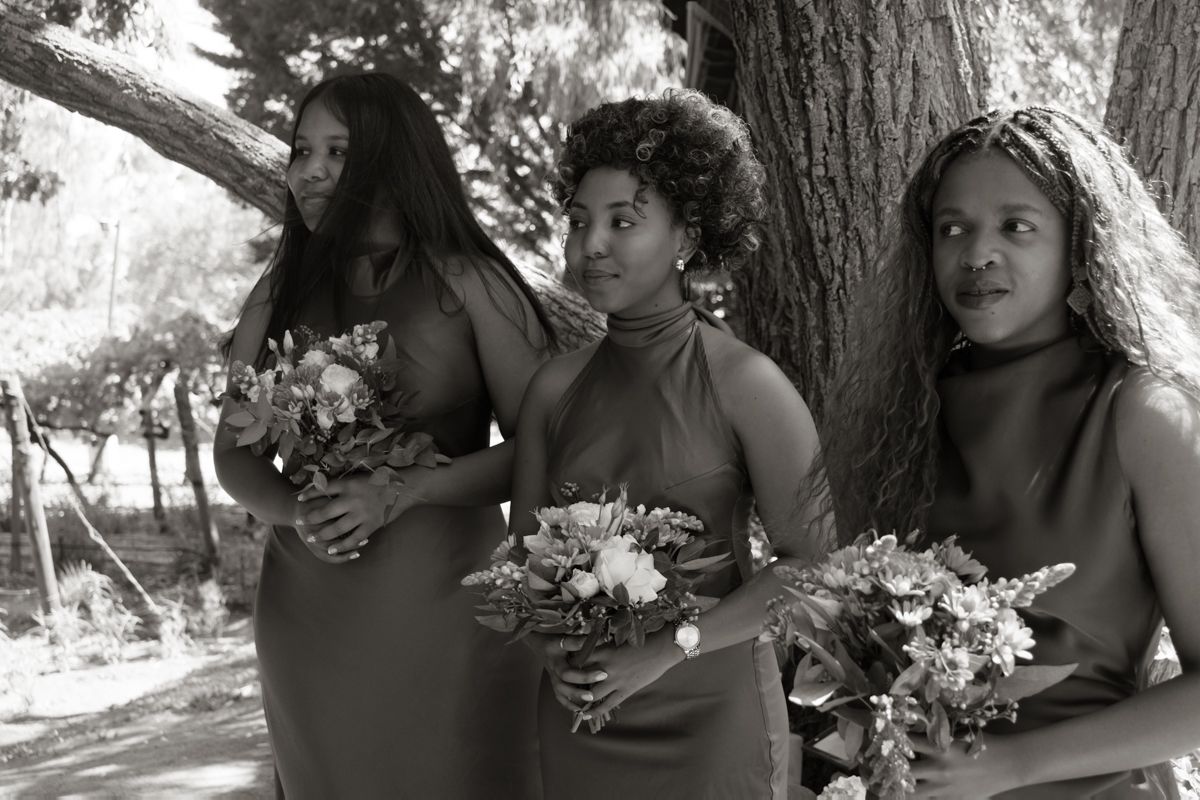 Three bridesmaids in halter dresses holding floral bouquets standing under a tree.