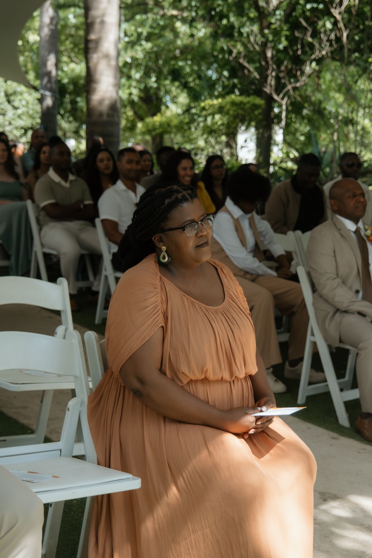 A woman in a peach dress with glasses sits attentively at an outdoor wedding ceremony among guests.