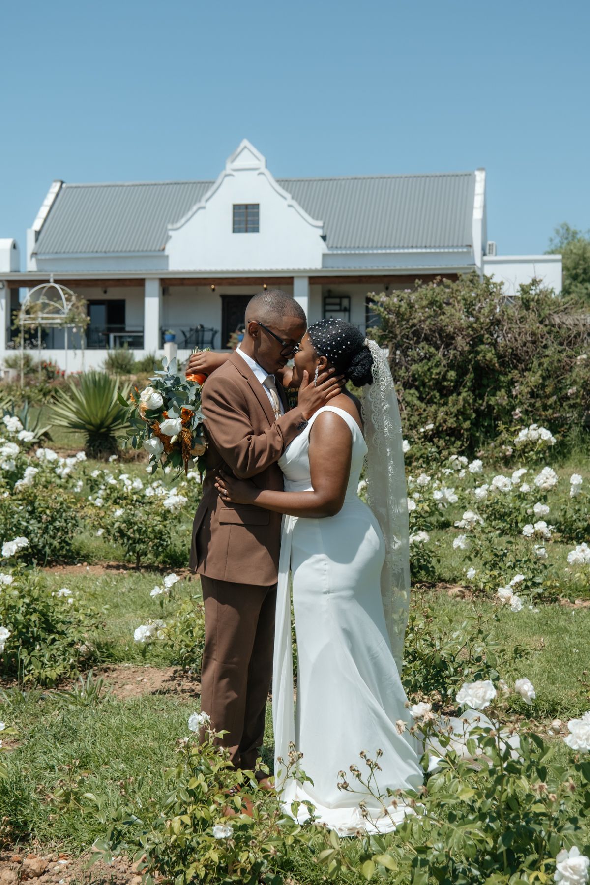 Black couple sharing a kiss in a rose garden with a white Cape Dutch wedding venue in the background.