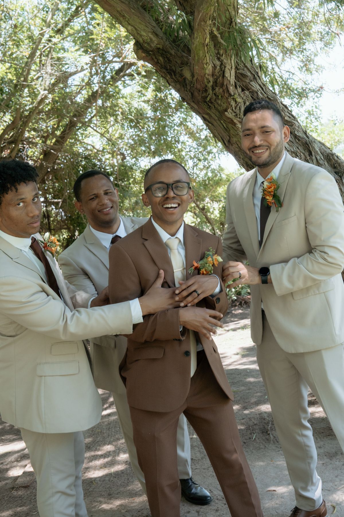 Groom in brown suit laughing with three groomsmen in beige suits under a tree, adjusting boutonnieres outdoors.
