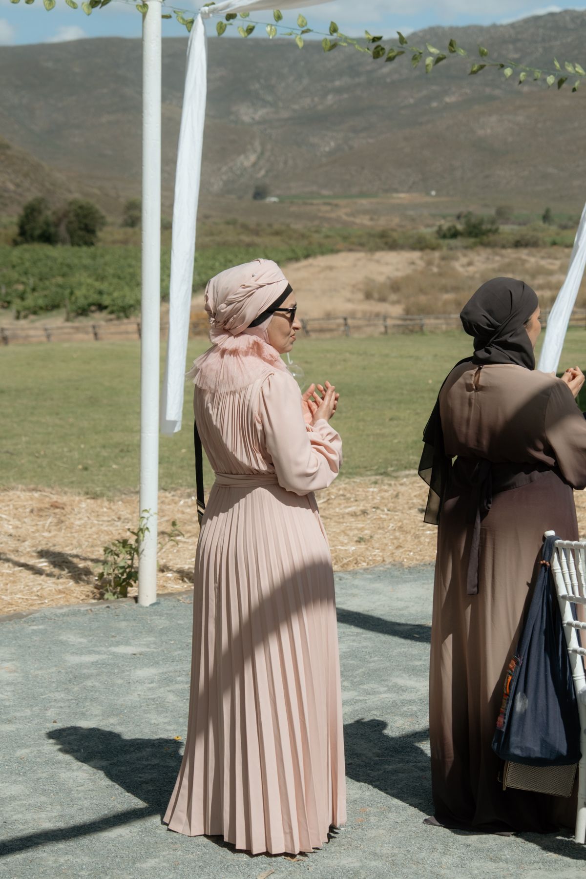 Two Muslim women in modest dresses and hijabs stand praying at outdoor wedding venue with mountain backdrop.