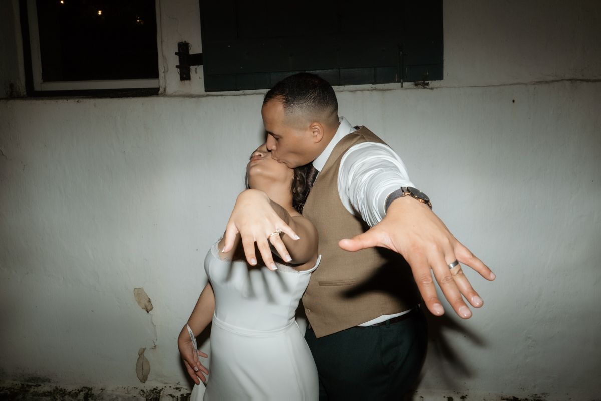 Groom dips bride for a kiss at wedding reception against a white wall in a candid romantic moment.