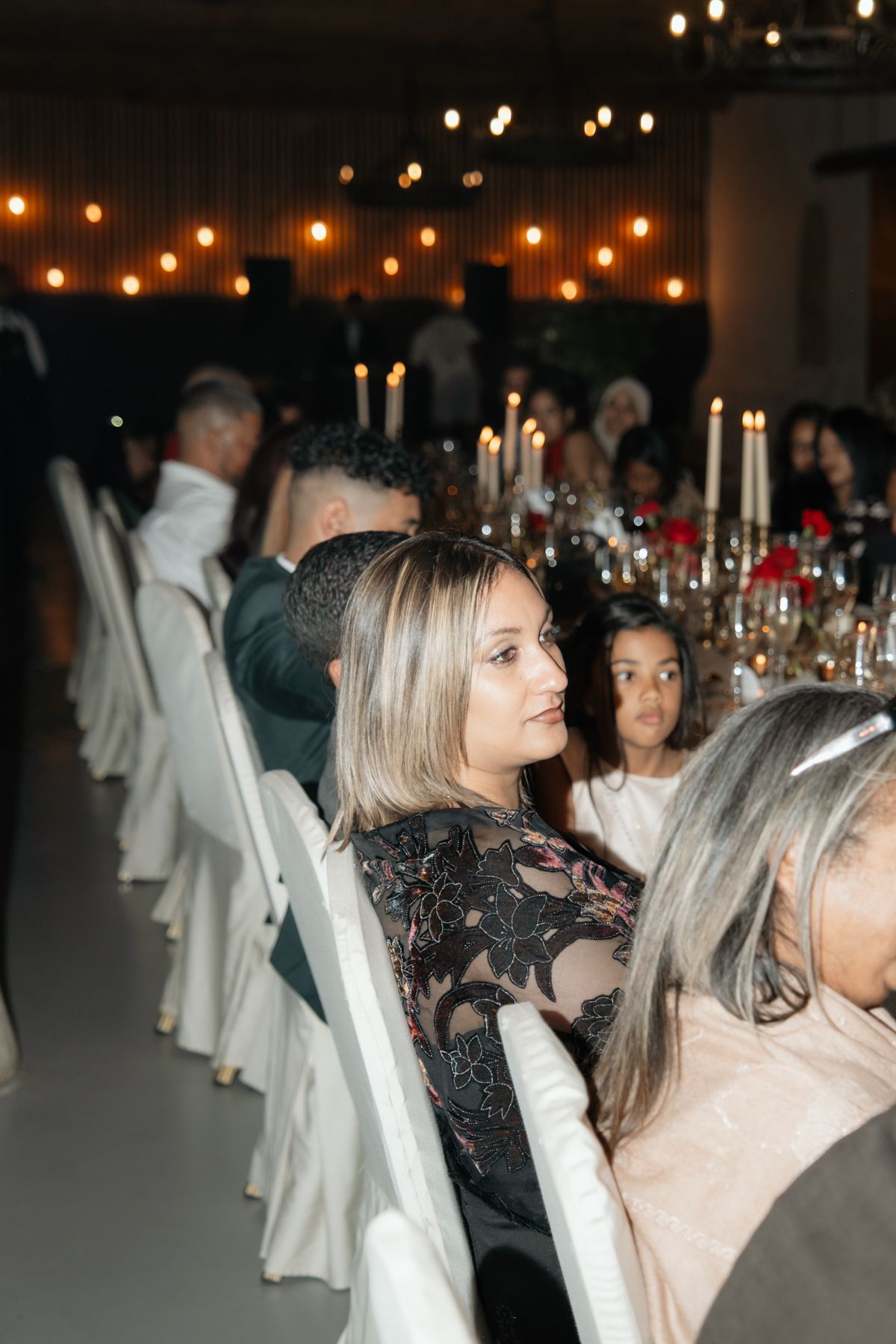 Wedding guest in black lace dress seated at candlelit reception table with string lights glowing overhead.