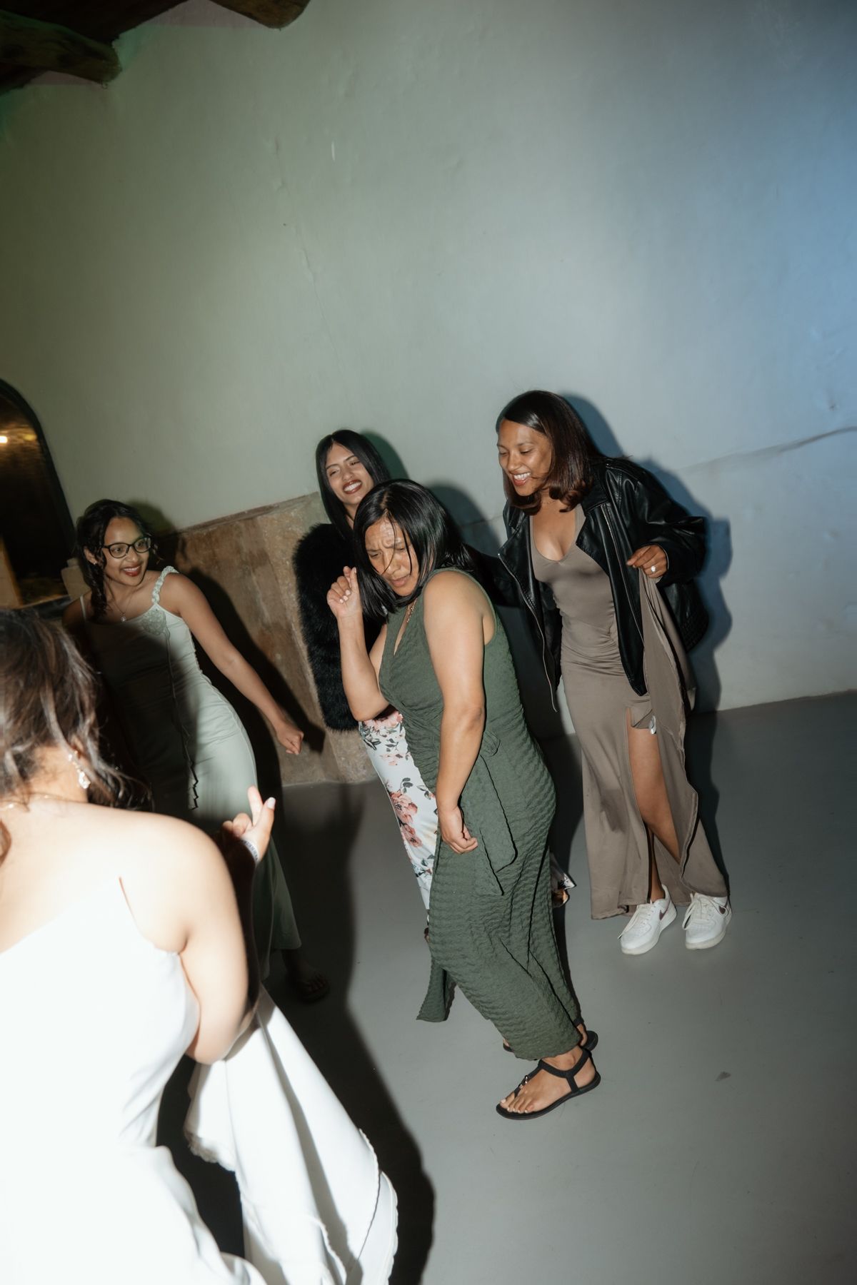 Group of women in elegant dresses dancing and laughing together at a wedding reception.