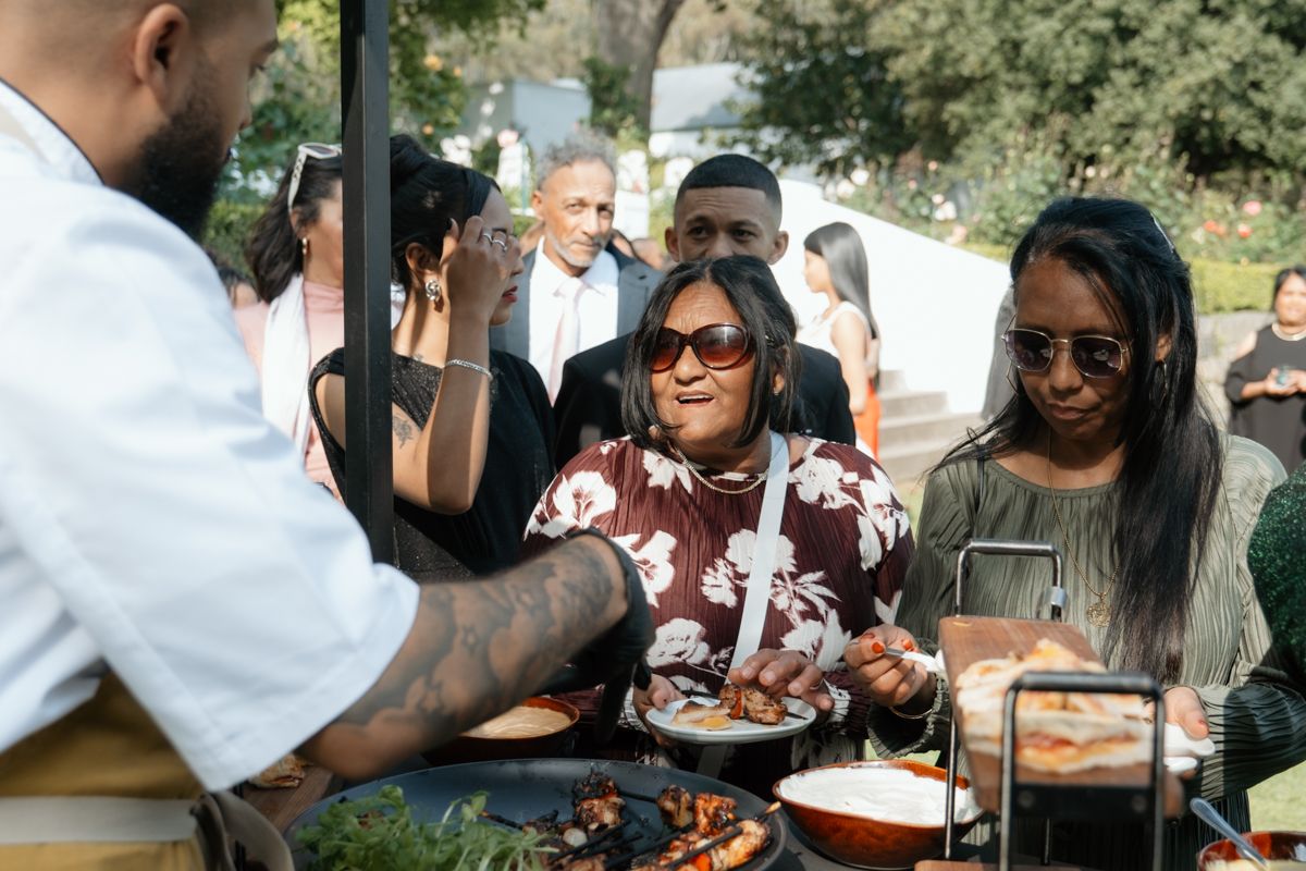 Wedding guests enjoying outdoor food station with chef serving grilled dishes at reception.