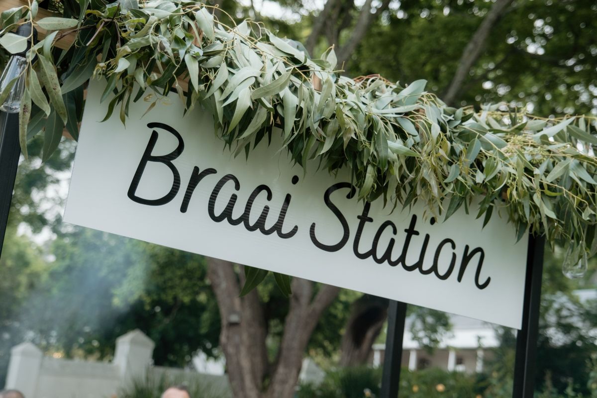 A decorative Braai Station sign adorned with olive branches at an outdoor wedding venue.