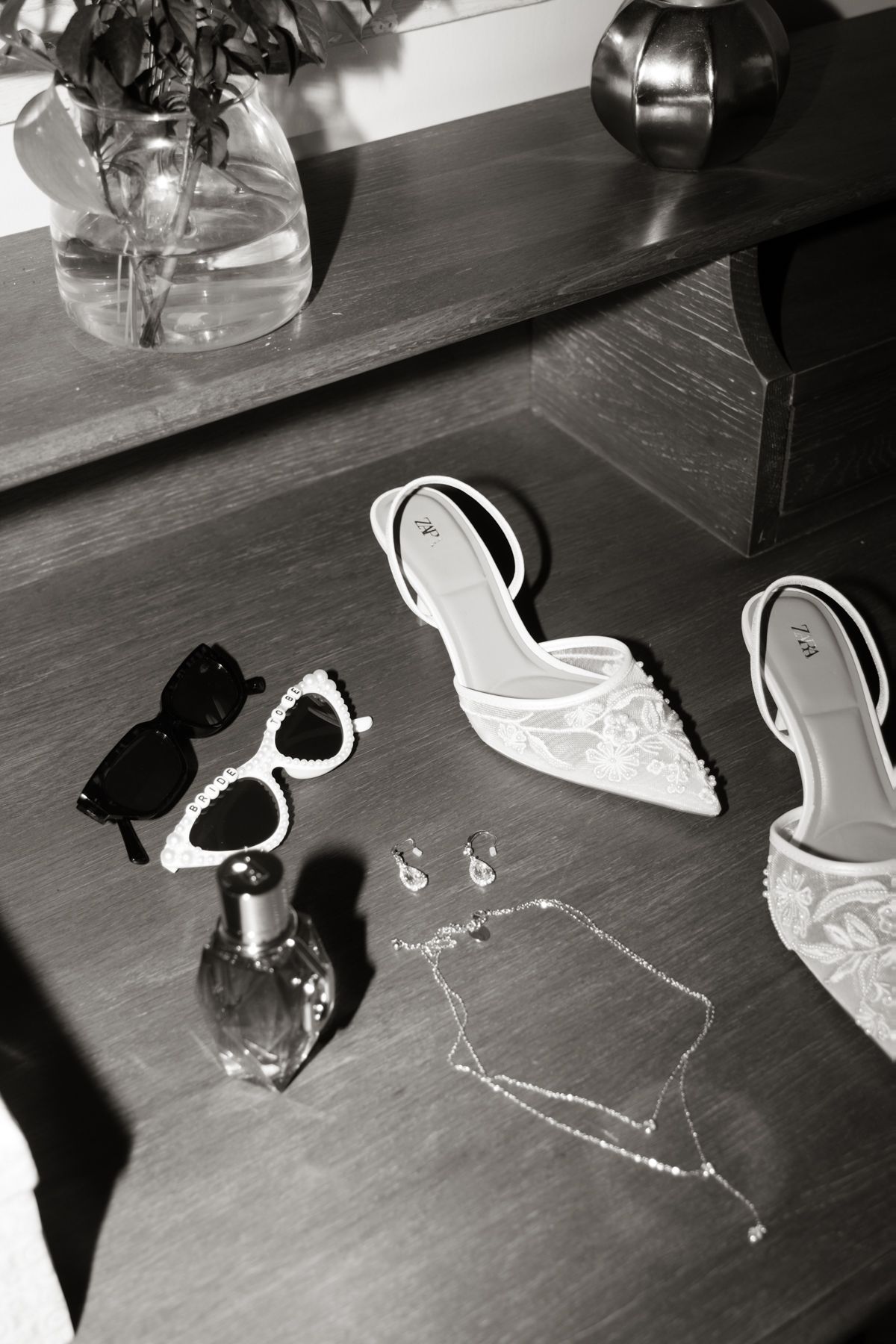 Black and white flat lay of bridal accessories including heels, sunglasses, perfume, earrings, and a delicate necklace.