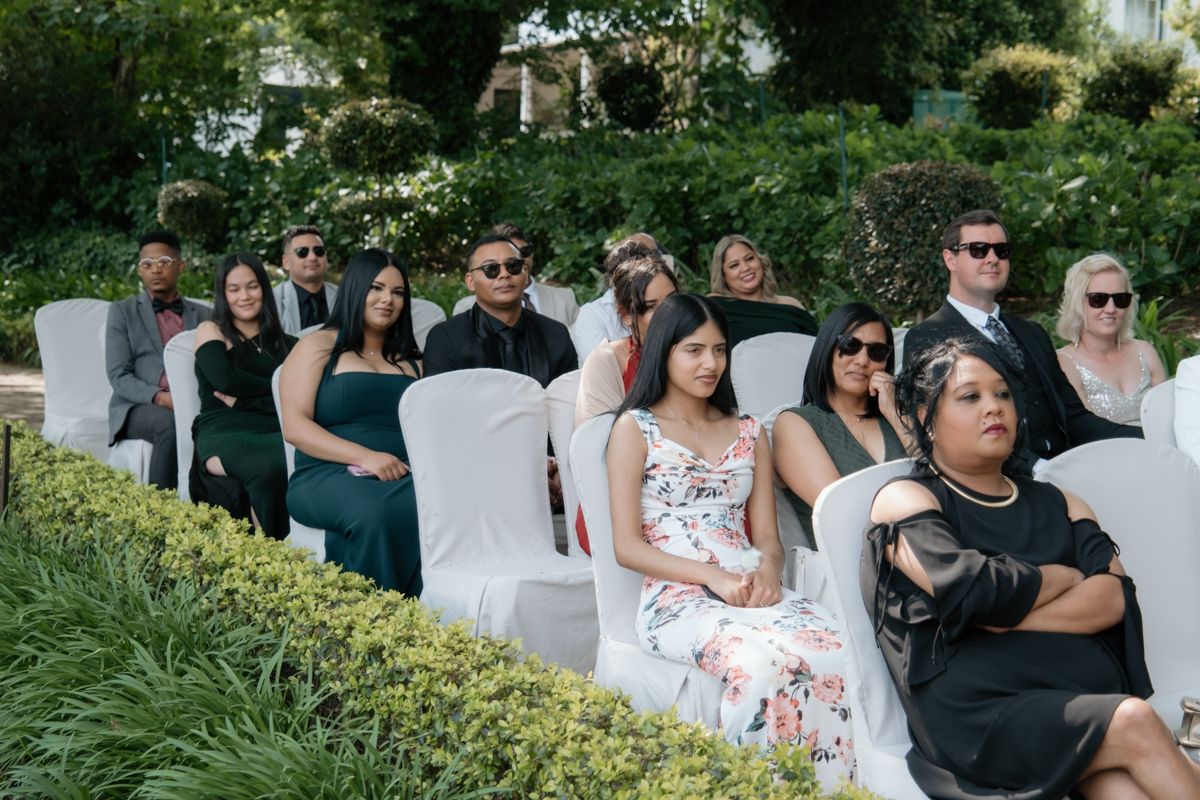 Wedding guests seated in white chairs outdoors watching ceremony in sunlit garden setting.