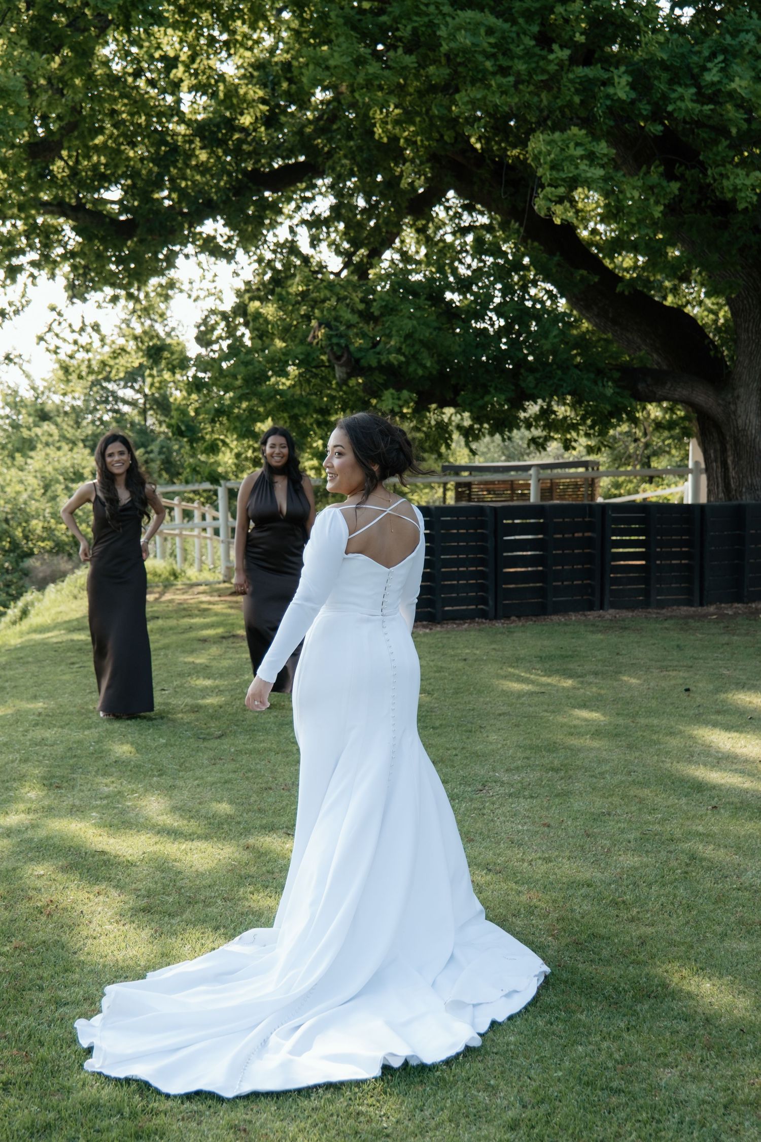 Smiling bride in white long-sleeve gown poses outdoors with bridesmaids in black dresses.