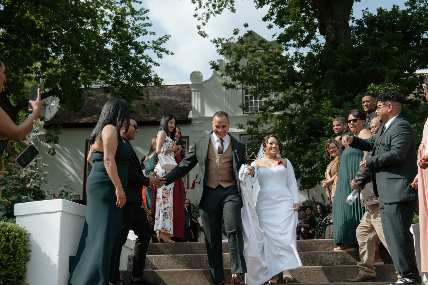 Newlyweds walk down steps surrounded by cheering wedding guests at outdoor venue.