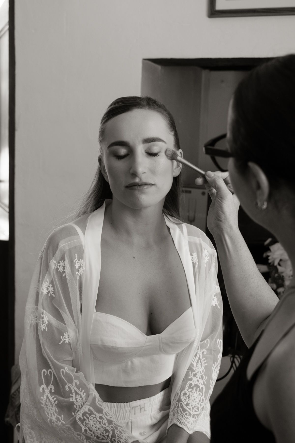 Black and white photo of a bride having makeup applied by a makeup artist while wearing a lace robe.
