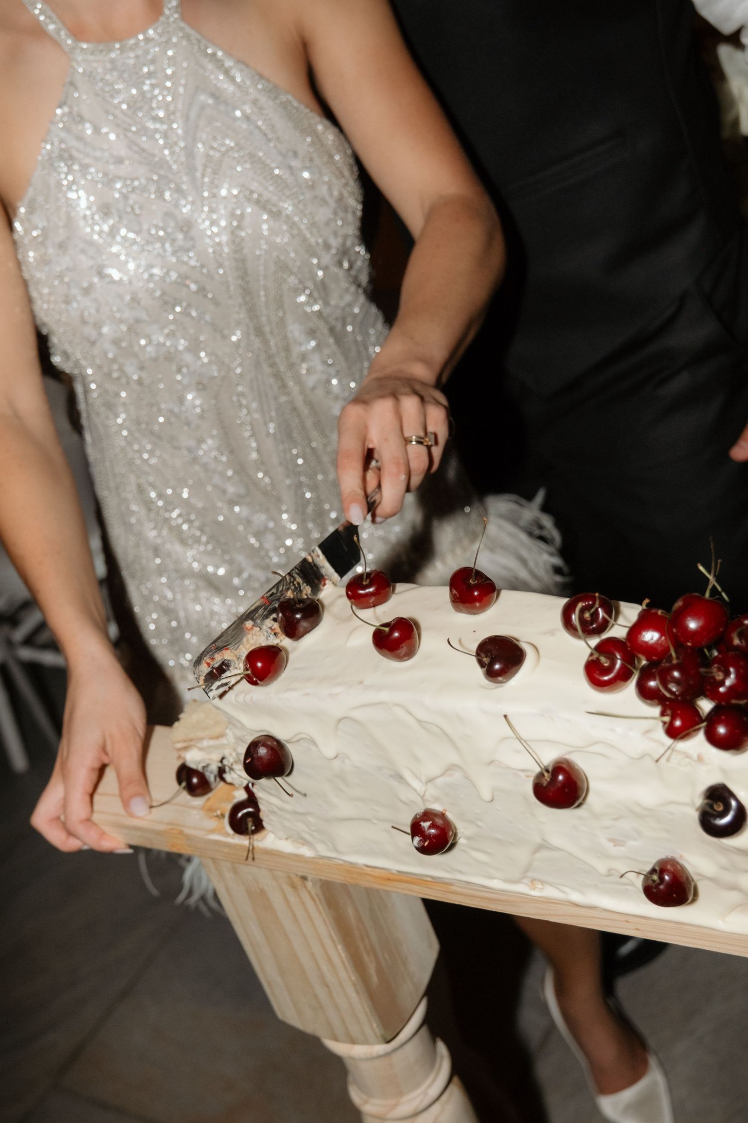 Couple cutting rustic wedding cake topped with fresh cherries, bride wearing silver sequin reception dress.