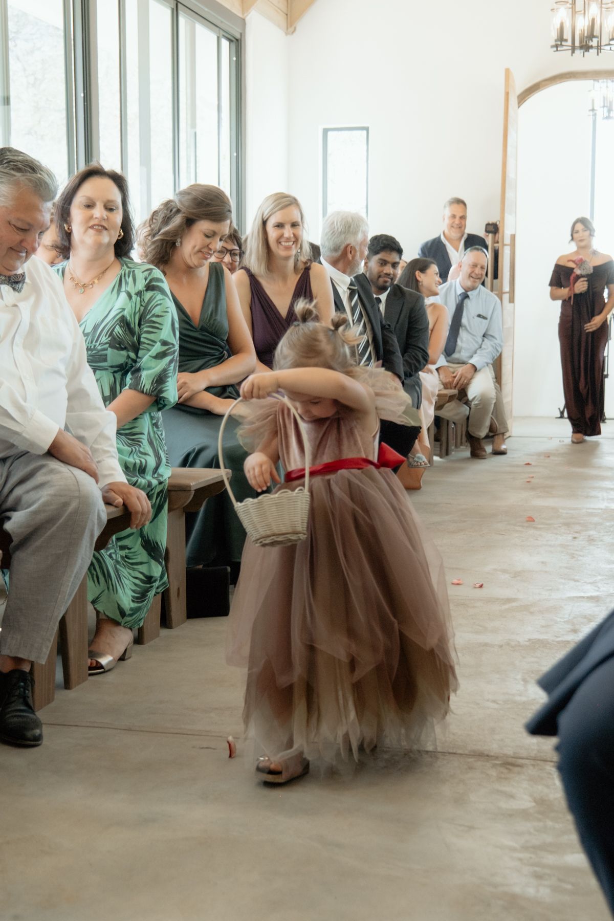 Flower girl in tulle dress walks down wedding aisle carrying basket while guests smile and watch.