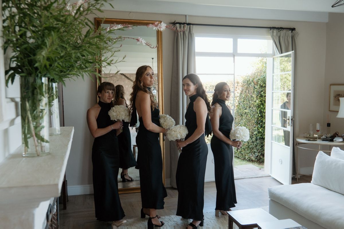 Four bridesmaids in elegant black dresses hold white bouquets while gathered in a bright, modern room.