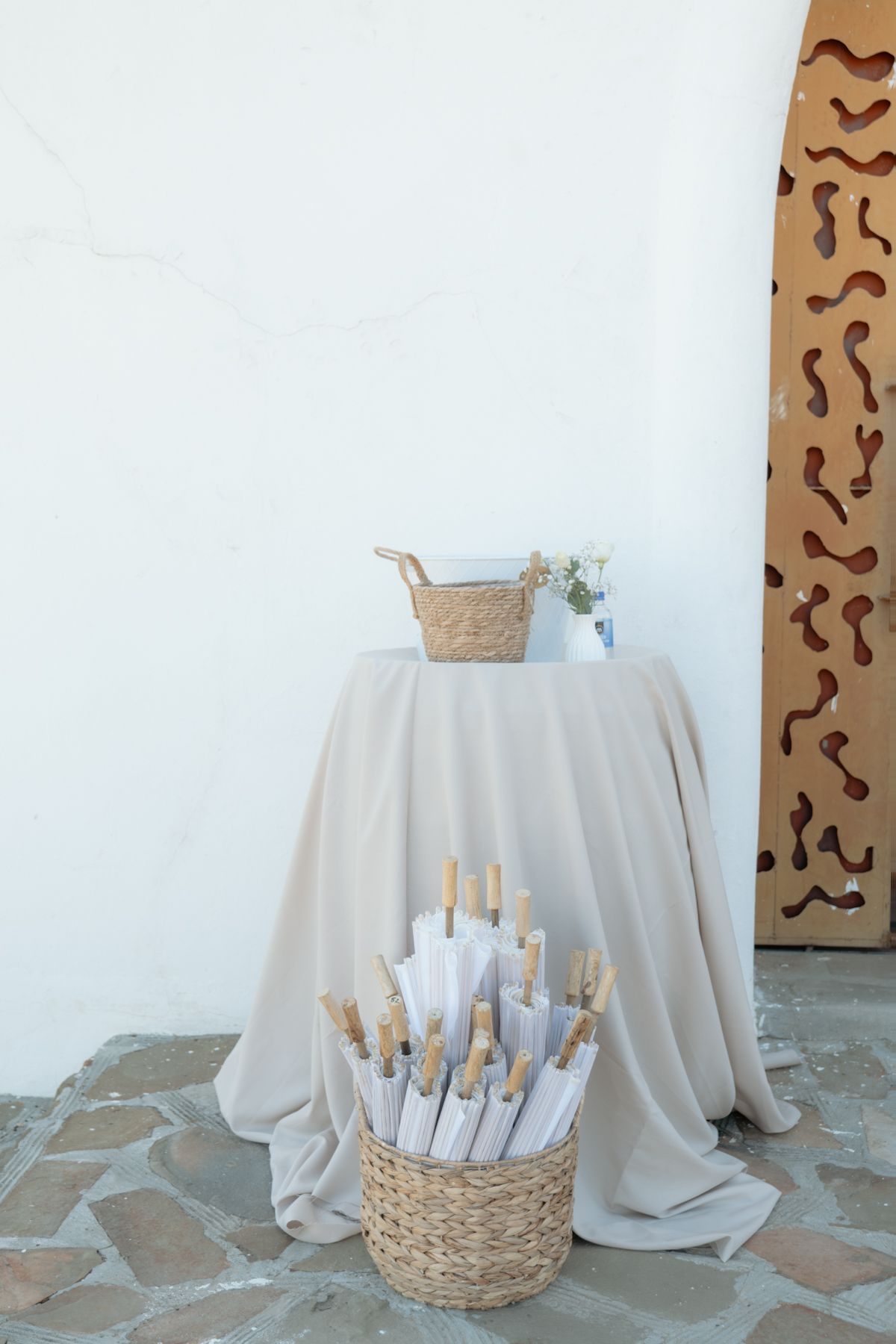 White parasols in a wicker basket beside a draped table at an outdoor wedding venue entrance.