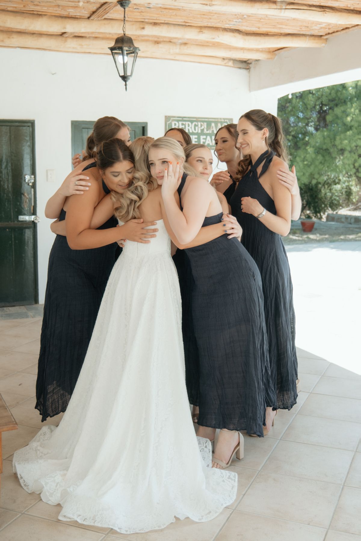 Bride in white gown embraced by bridesmaids in dark dresses at a rustic wedding venue.