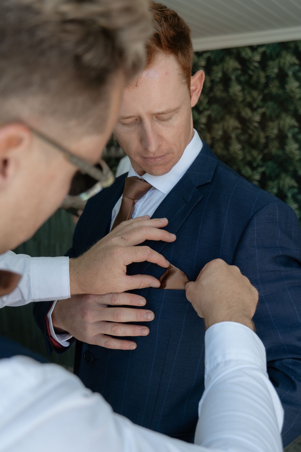 Groomsman adjusting boutonniere on groom wearing navy suit and brown tie before wedding ceremony outdoors.