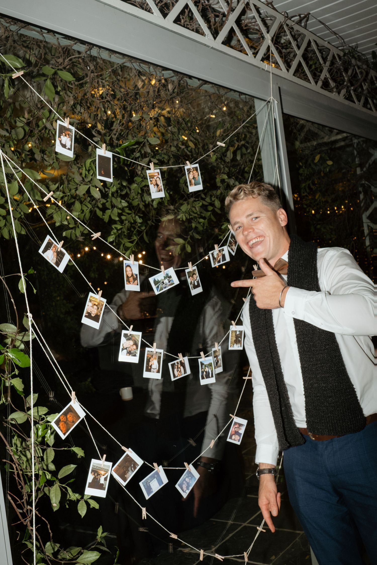 Wedding guest pointing at Polaroid photo display strung with fairy lights at evening reception.