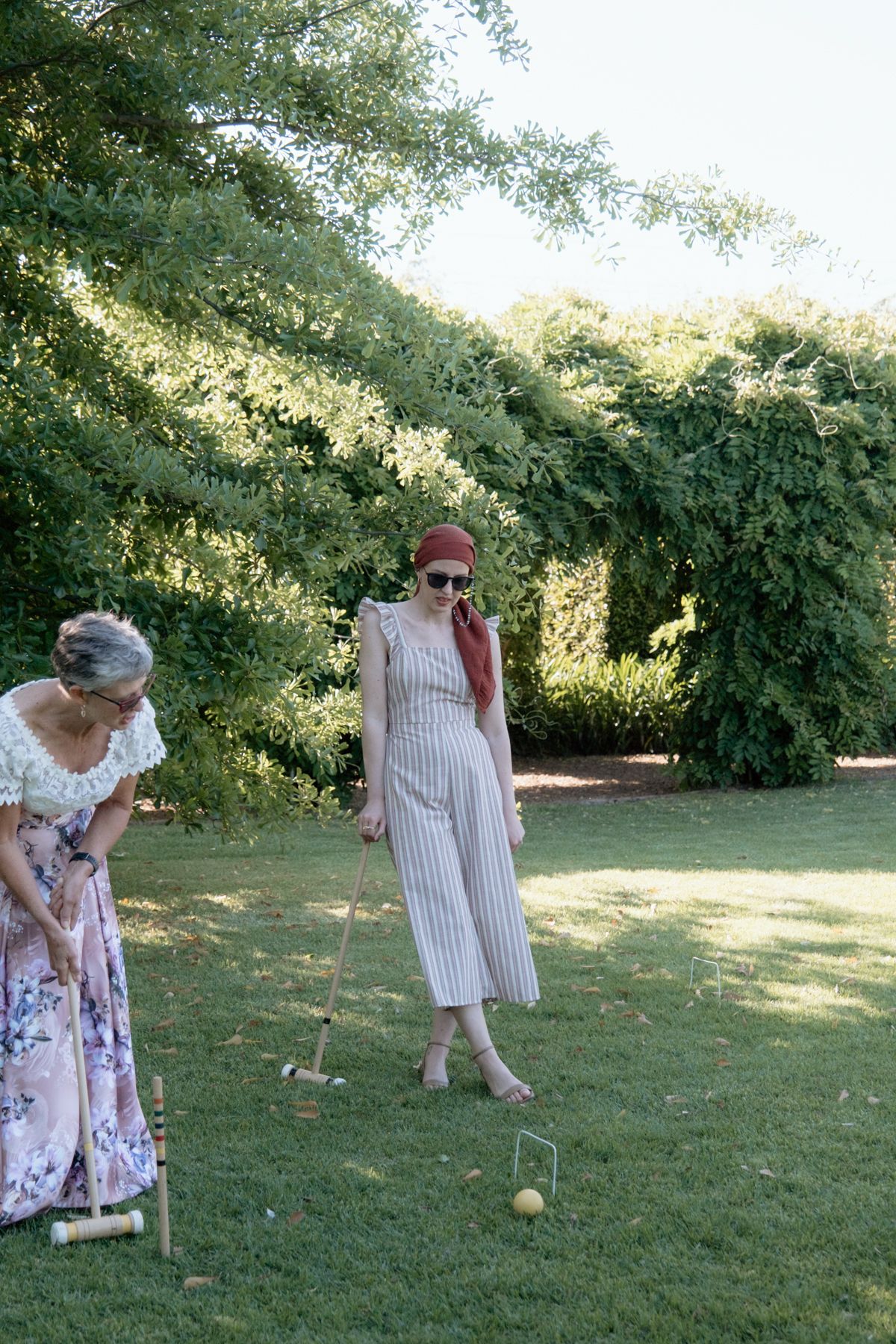 Two women play croquet on a lush green lawn surrounded by trees at a summer wedding reception.