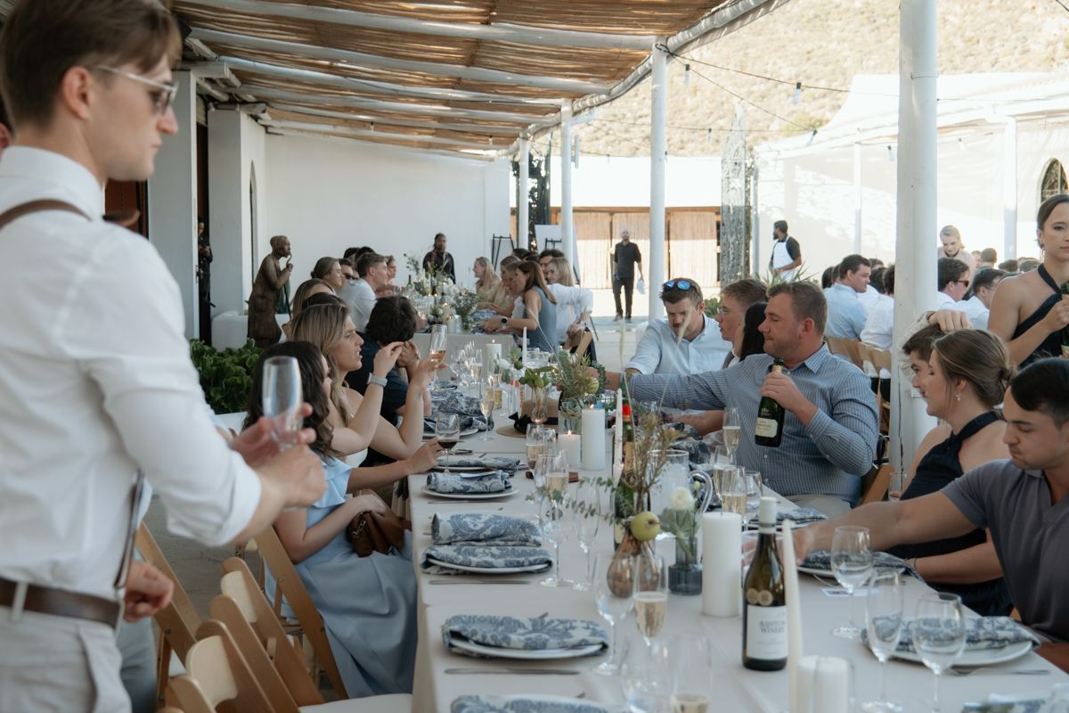 Wedding guests seated at long decorated reception table under pergola, raising glasses in a toast.