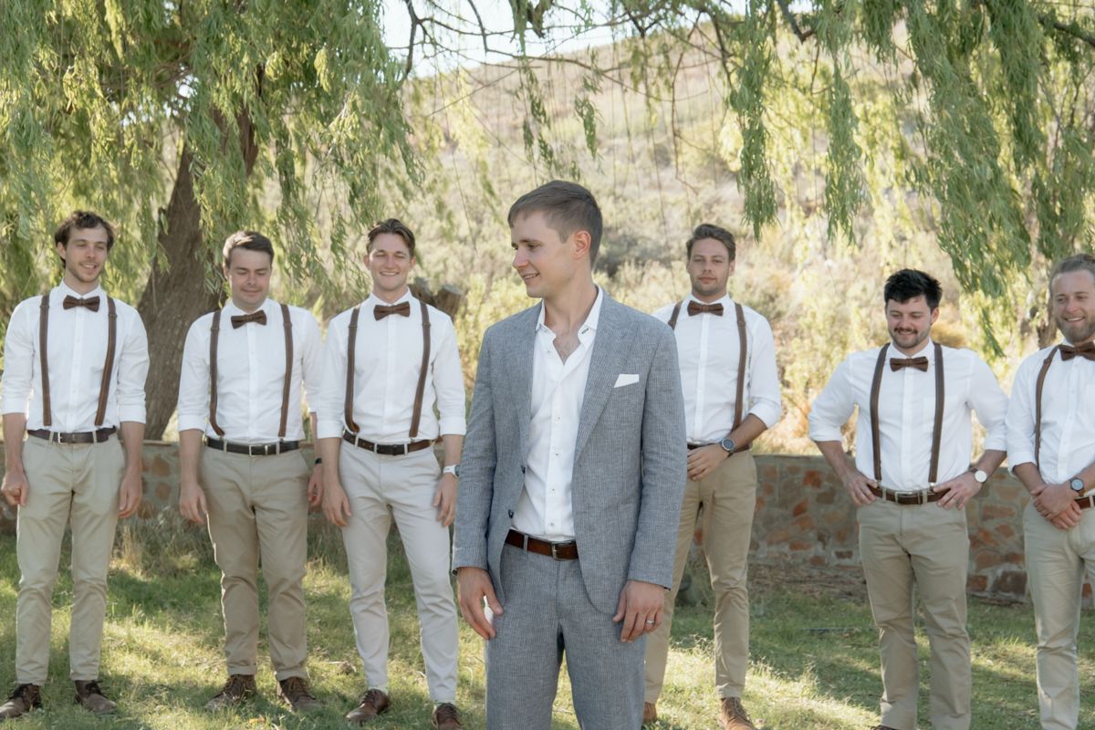 Groom in grey suit stands with groomsmen wearing suspenders and bow ties under willow tree.
