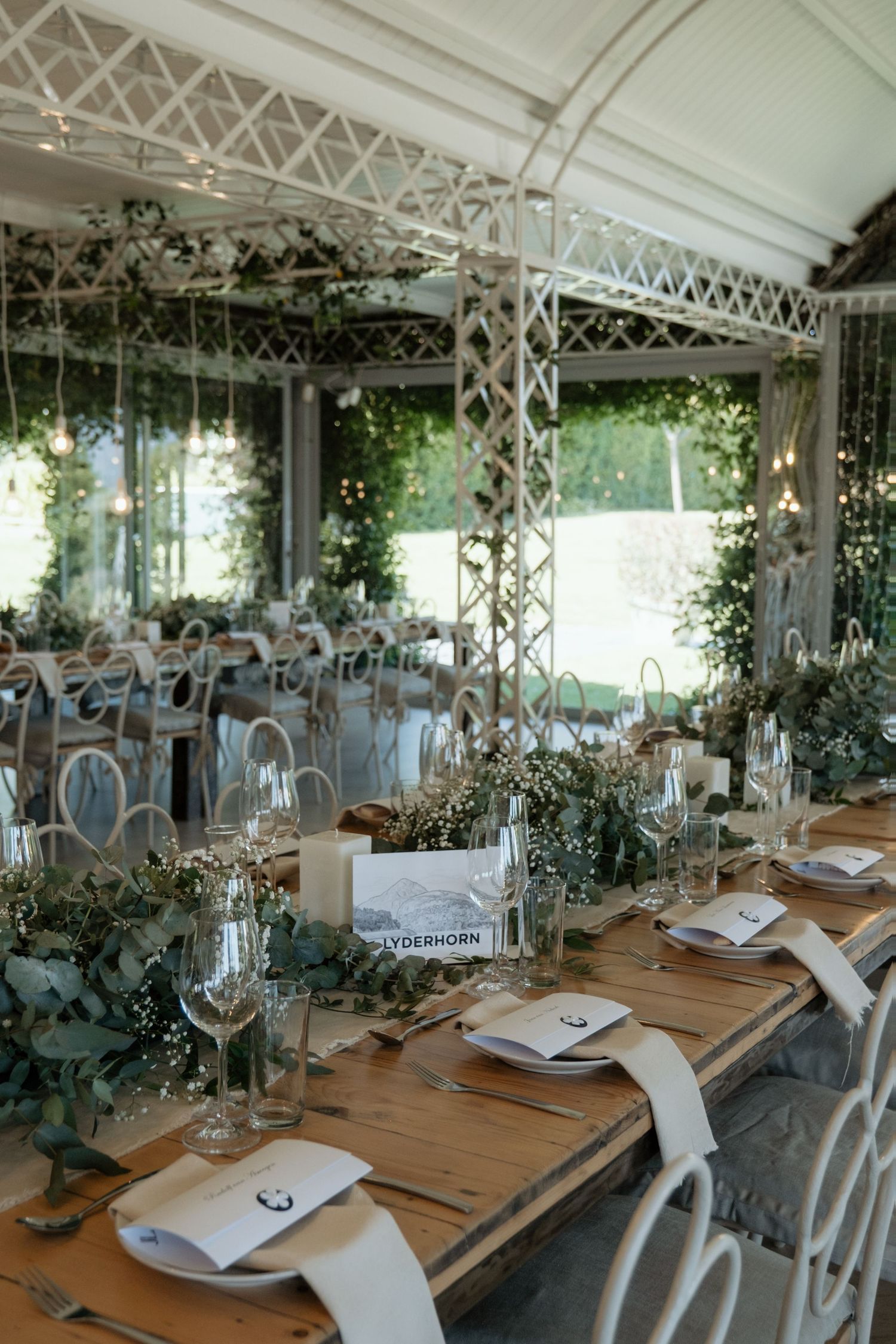Elegant wedding reception table with eucalyptus greenery runner, candles, and white chairs in a garden pavilion.