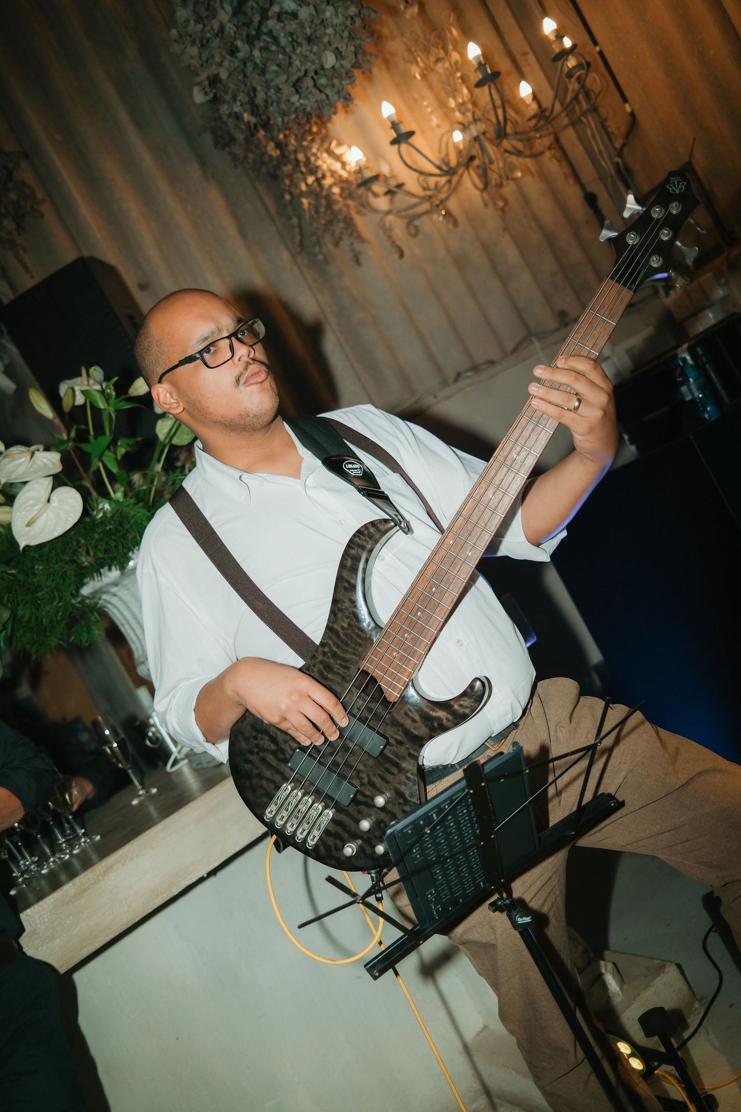 Bald musician in white shirt plays electric bass guitar at elegant wedding reception with chandelier backdrop.