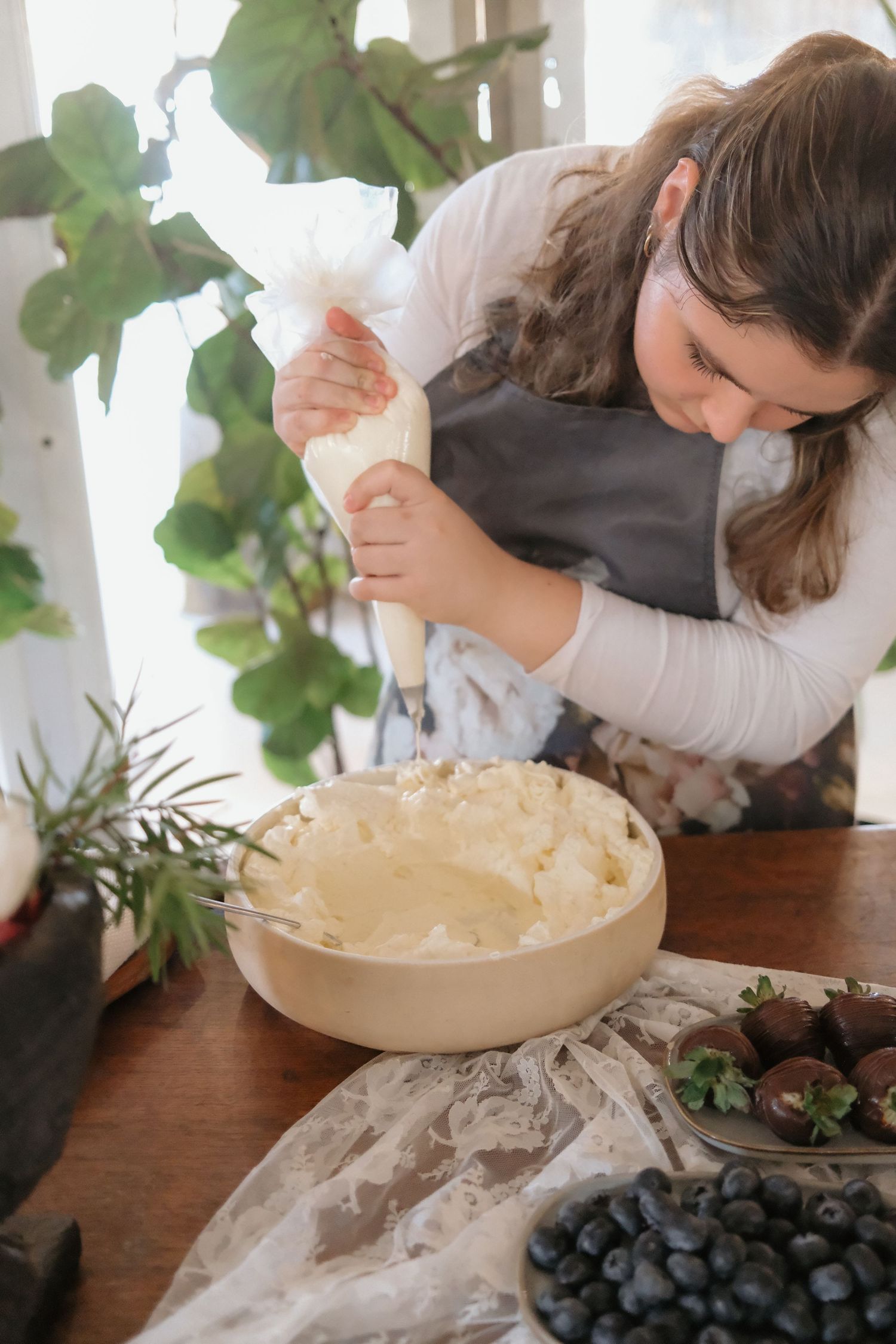 Baker pipes cream into a wooden bowl while preparing a dessert with blueberries and chocolate strawberries nearby.