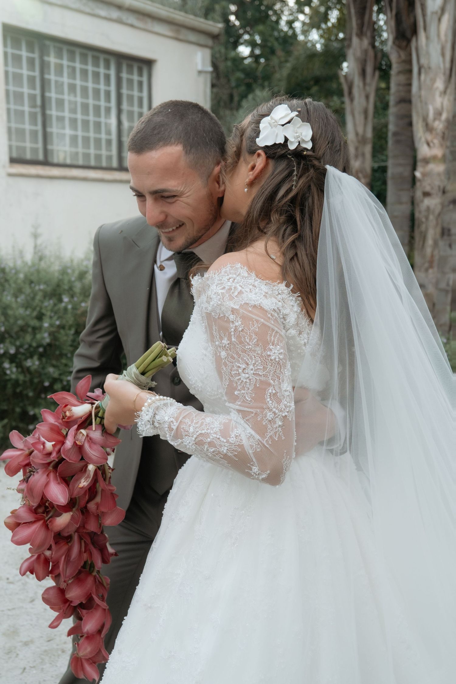 Smiling groom in gray suit with bride in lace wedding dress holding red floral bouquet outdoors.