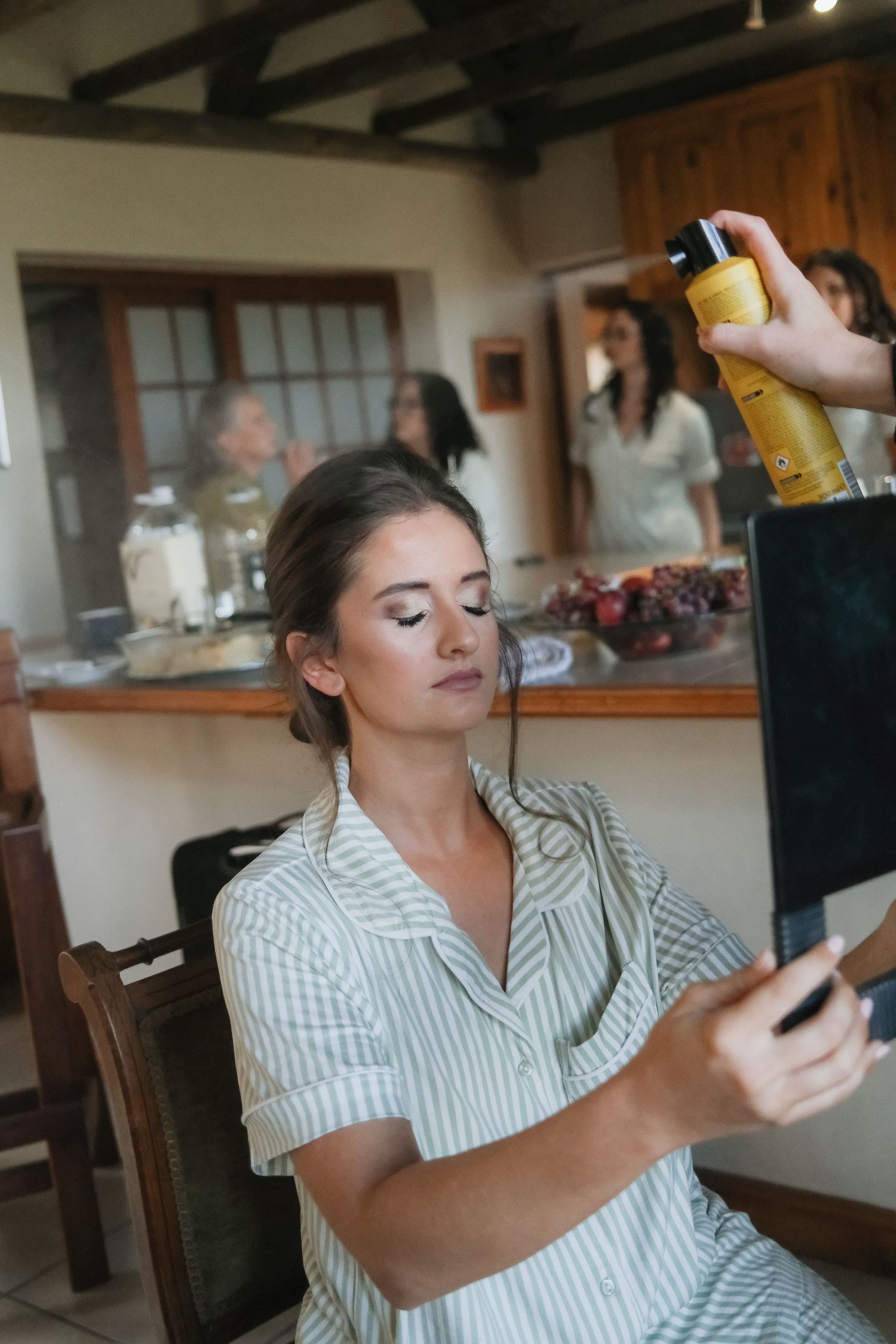 Bride in striped pajamas checks mirror while makeup artist applies hairspray during wedding prep.