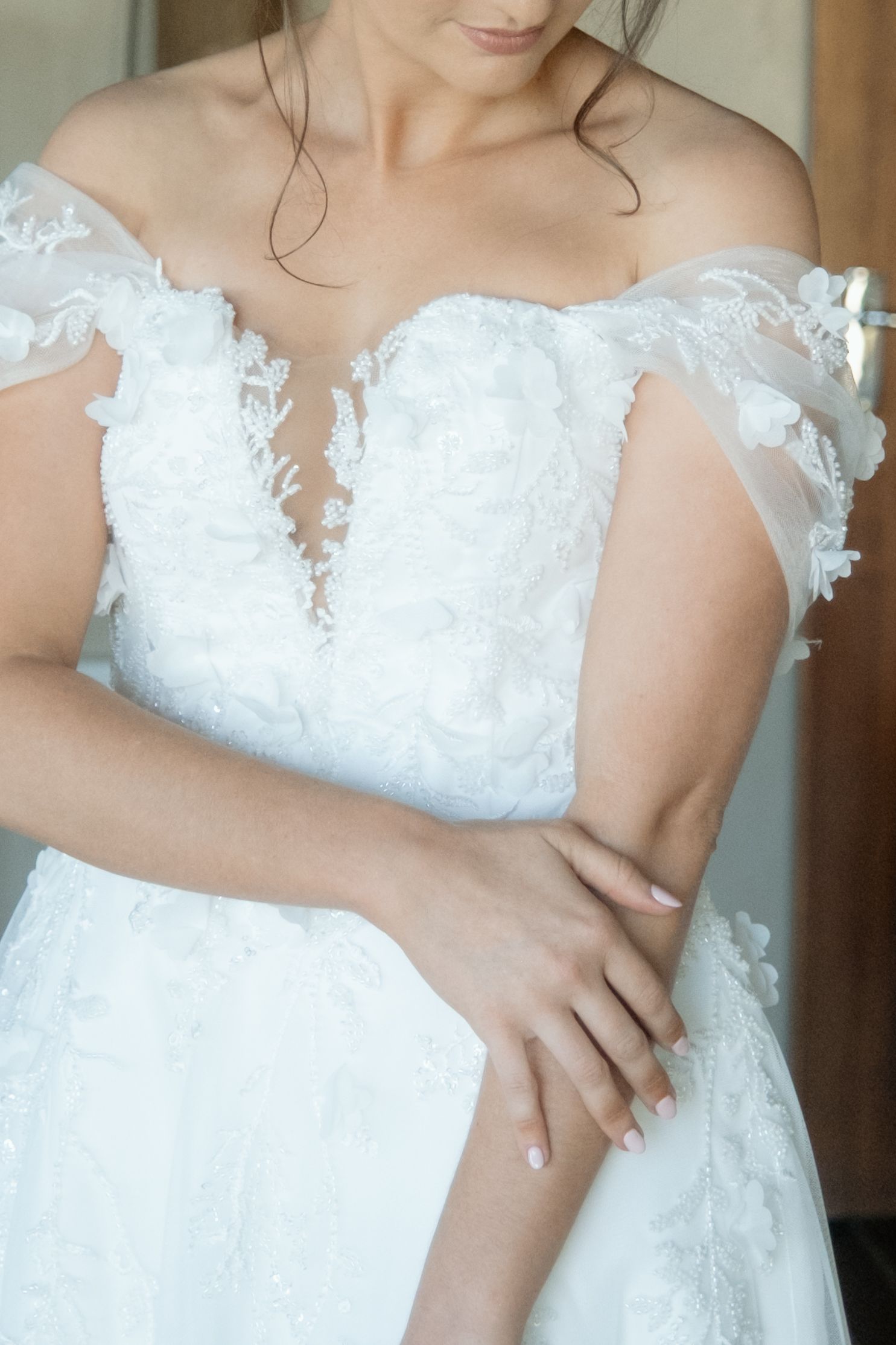 Bride in off-shoulder white lace wedding gown with floral appliques and sweetheart neckline, hands clasped at waist.